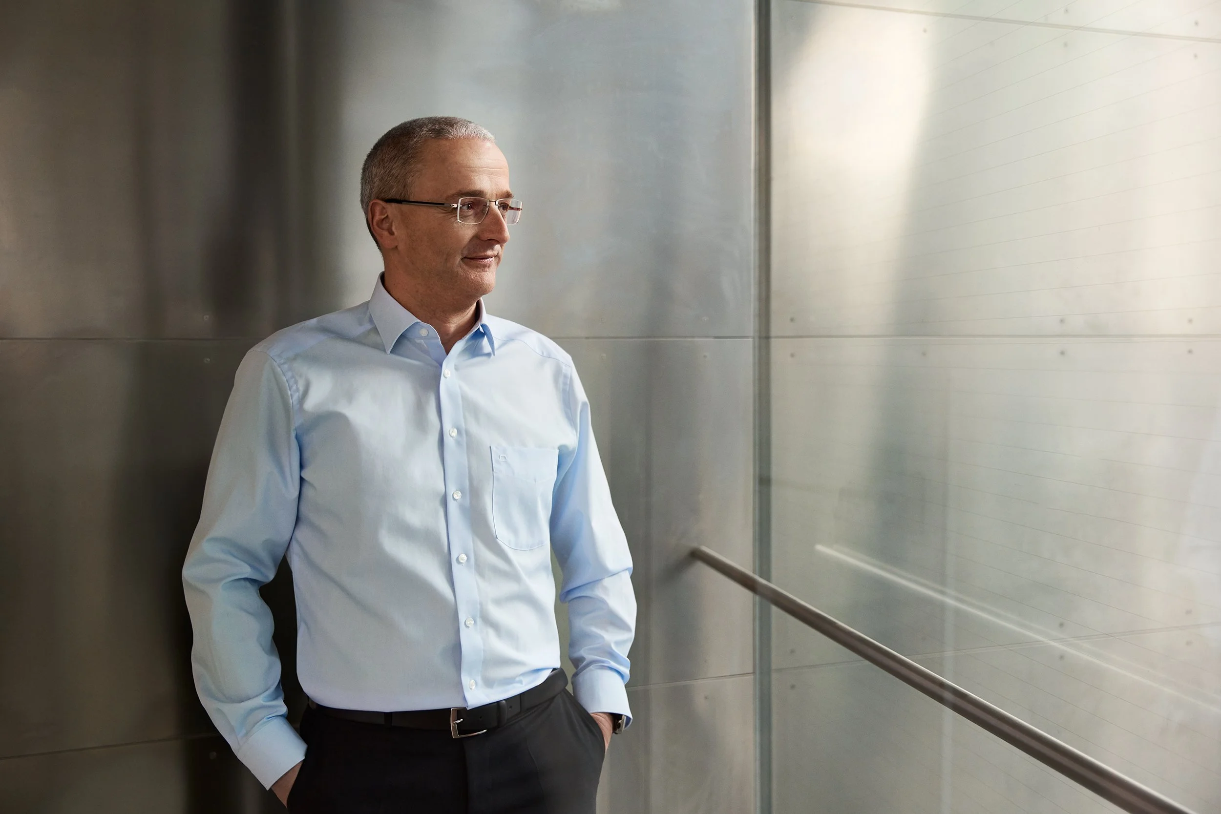 Ein Mann in Business-Kleidung steht in einem modernen, hell erleuchteten Flur mit metallem und sandgestrahltem Oberfläche, er lehnt an der Wand und blickt nach draußen.