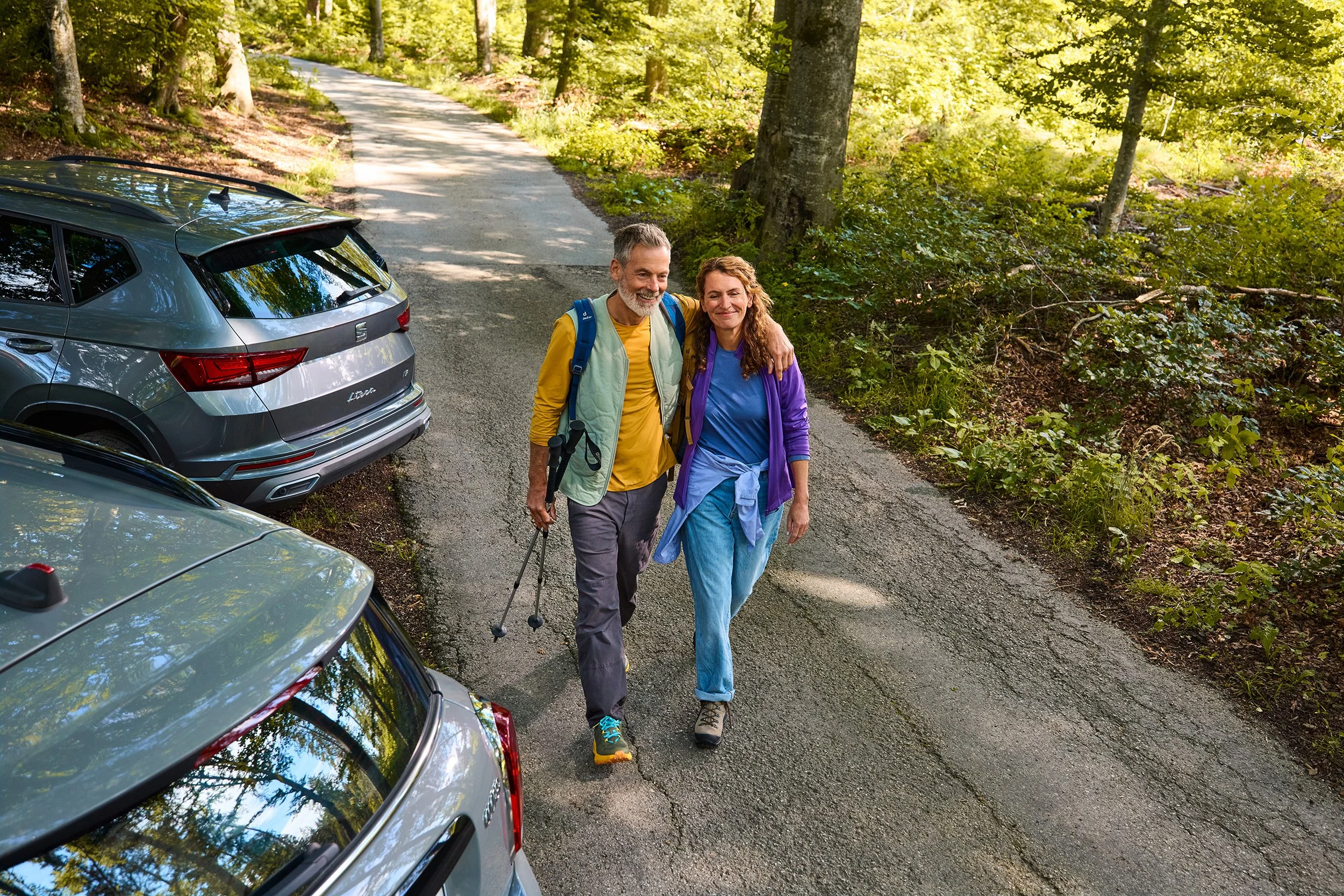 Ein älteres Paar wandert auf einer Waldstraße, umgeben von Bäumen und grünem Laub, mit parkenden Autos am Straßenrand.