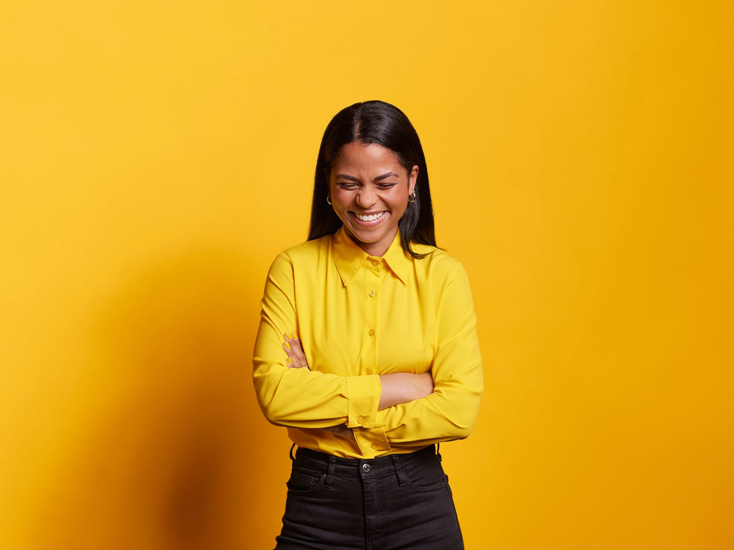 Junge Frau lachend vor gelbem Hintergrund, trägt gelbe Bluse und schwarze Hose.