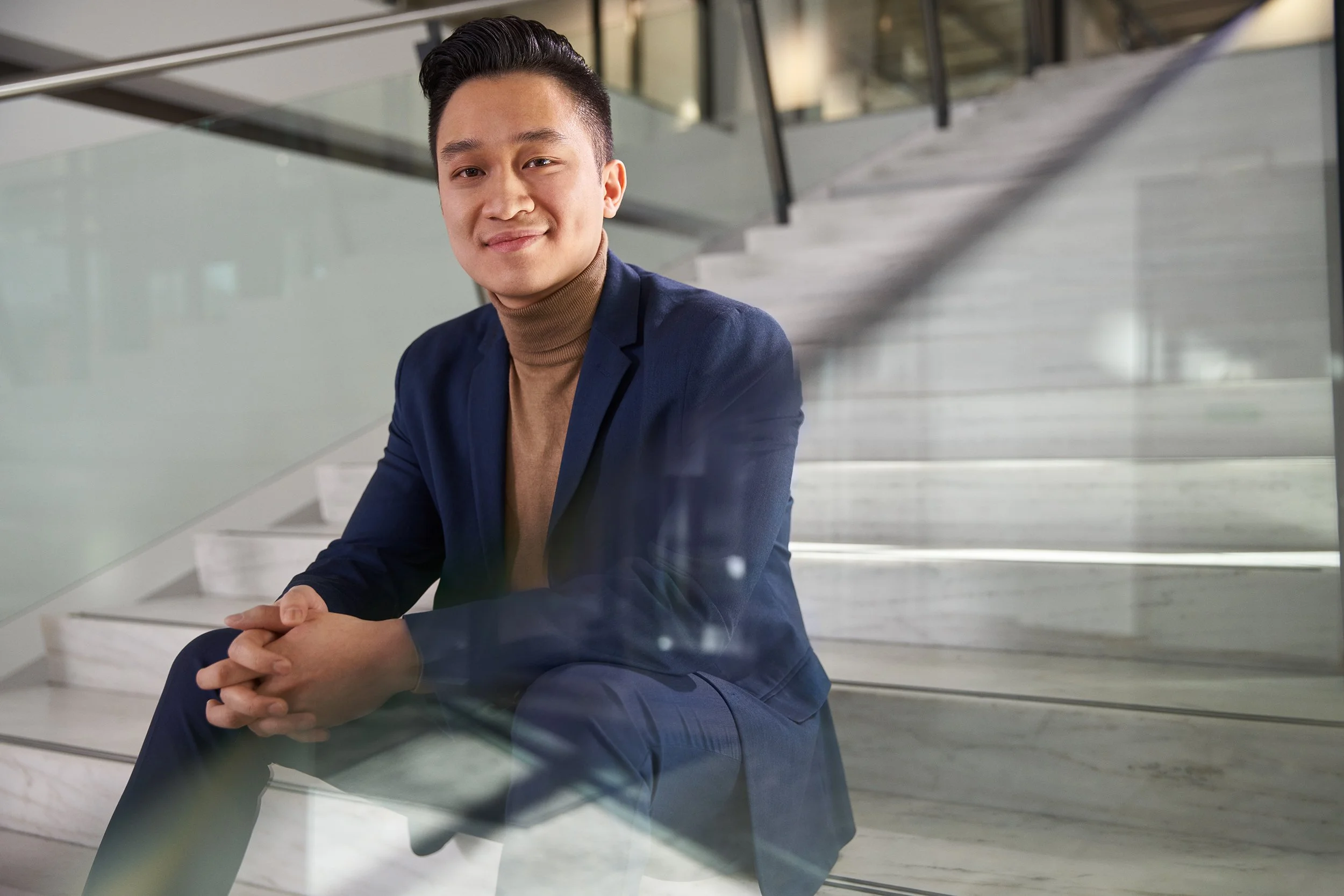 Ein junger Mann in einem blauen Anzug sitzt auf weißen Marmorstufen in einem modernen Gebäude, lächelt und sieht in die Kamera.