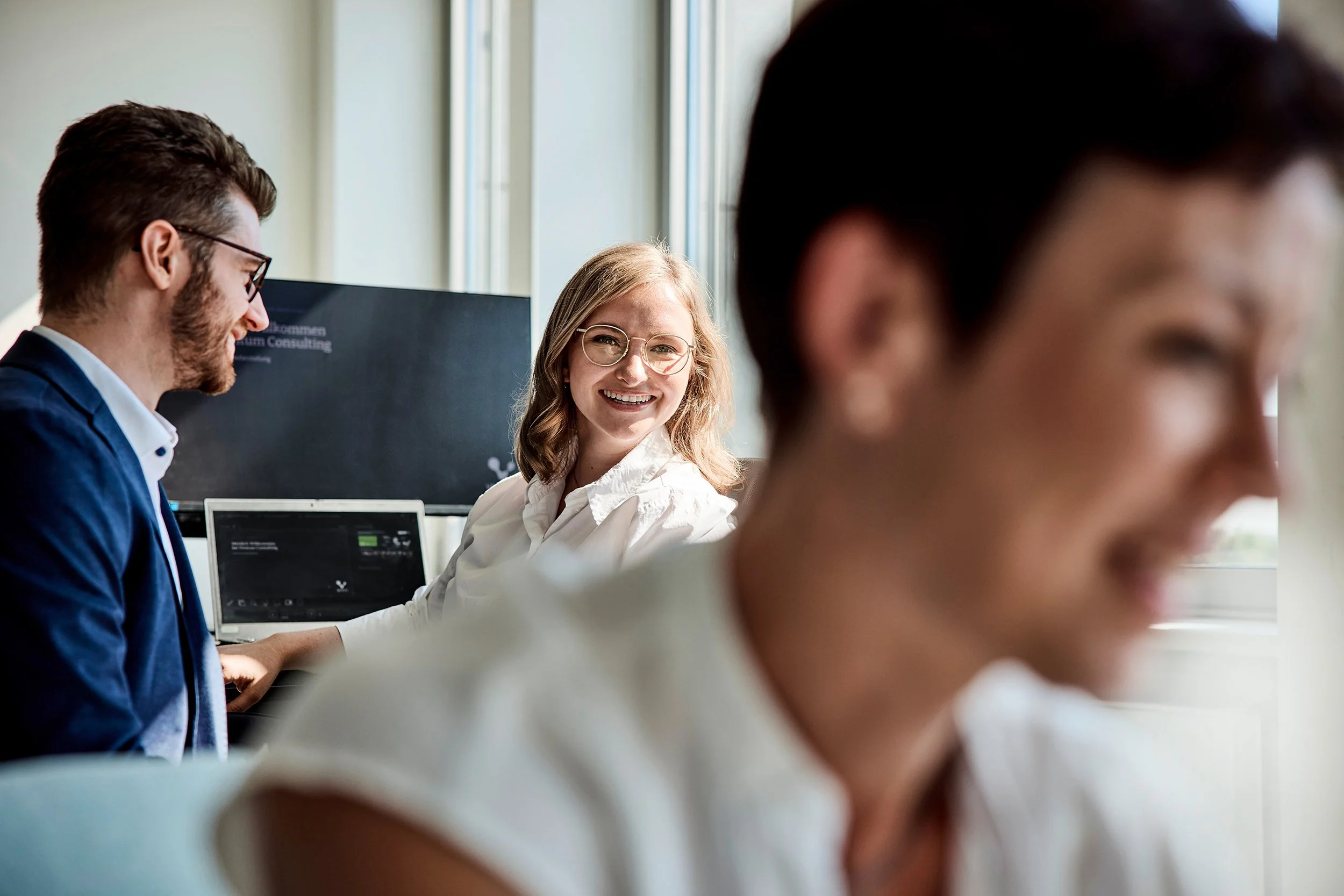 Drei junge Menschen lachen und unterhalten sich in einem Büro, im Hintergrund ein Computerbildschirm und ein weiterer Monitor.