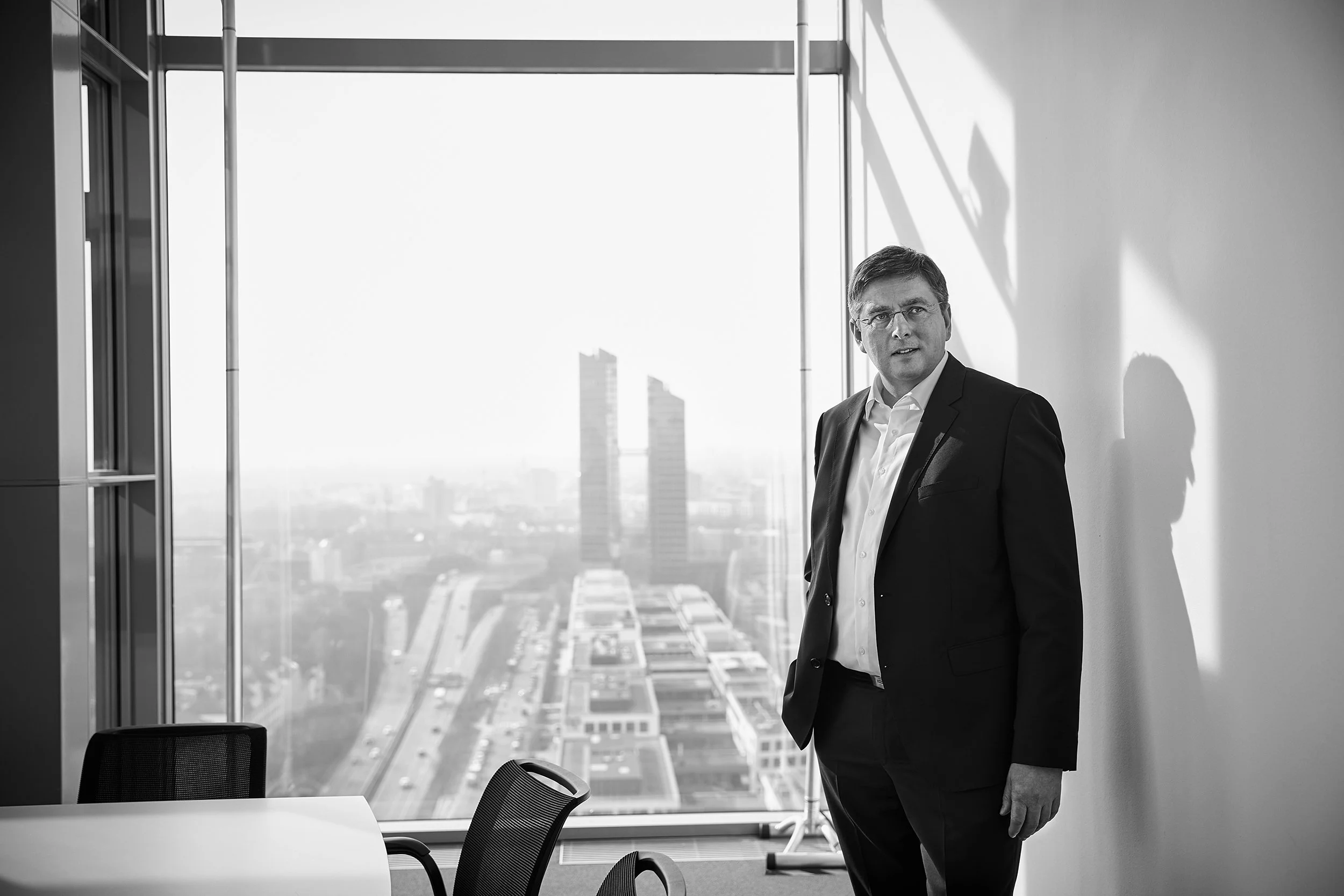 Mann in Anzug steht in einem modernen Büro mit Fensterblick auf eine Stadt. Deutlich sind ein Tisch und Stühle sichtbar.