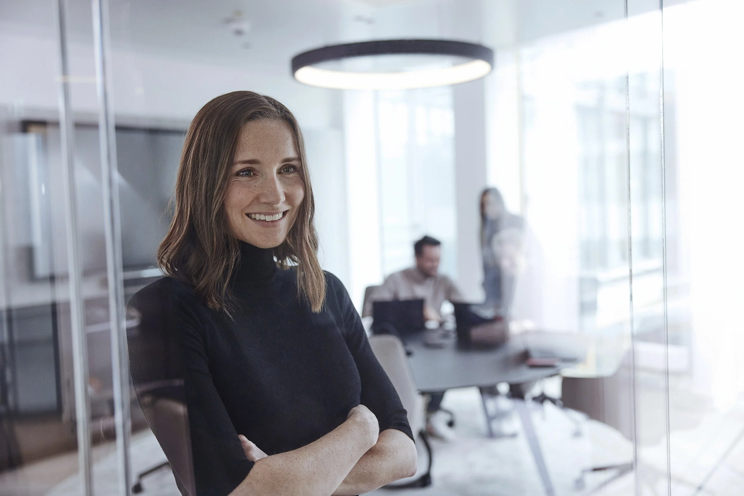 Eine lachende Frau in einem schwarzen Rollkragenpullover steht vor einem Glasfenster in einem modernen Büro, im Hintergrund arbeiten weitere Menschen an einem Tisch.