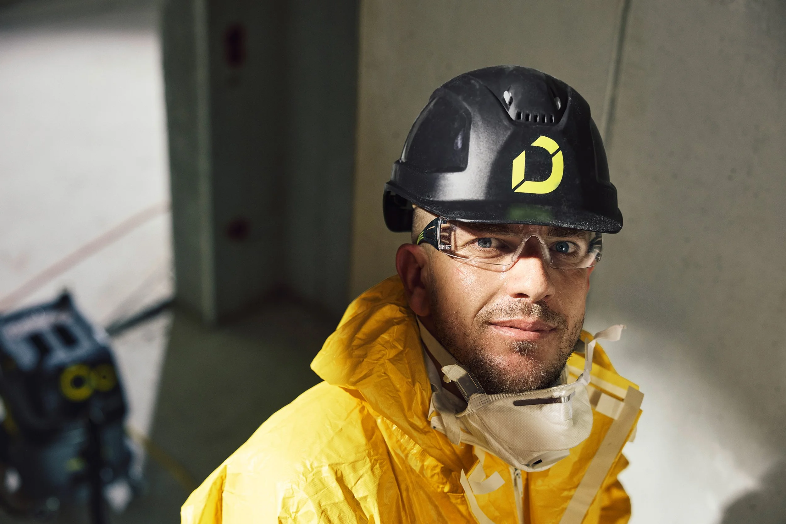 Bauarbeiter mit Schutzhelm und gelber Jacke, blickt in die Kamera, im Hintergrund Baustellenumgebung.