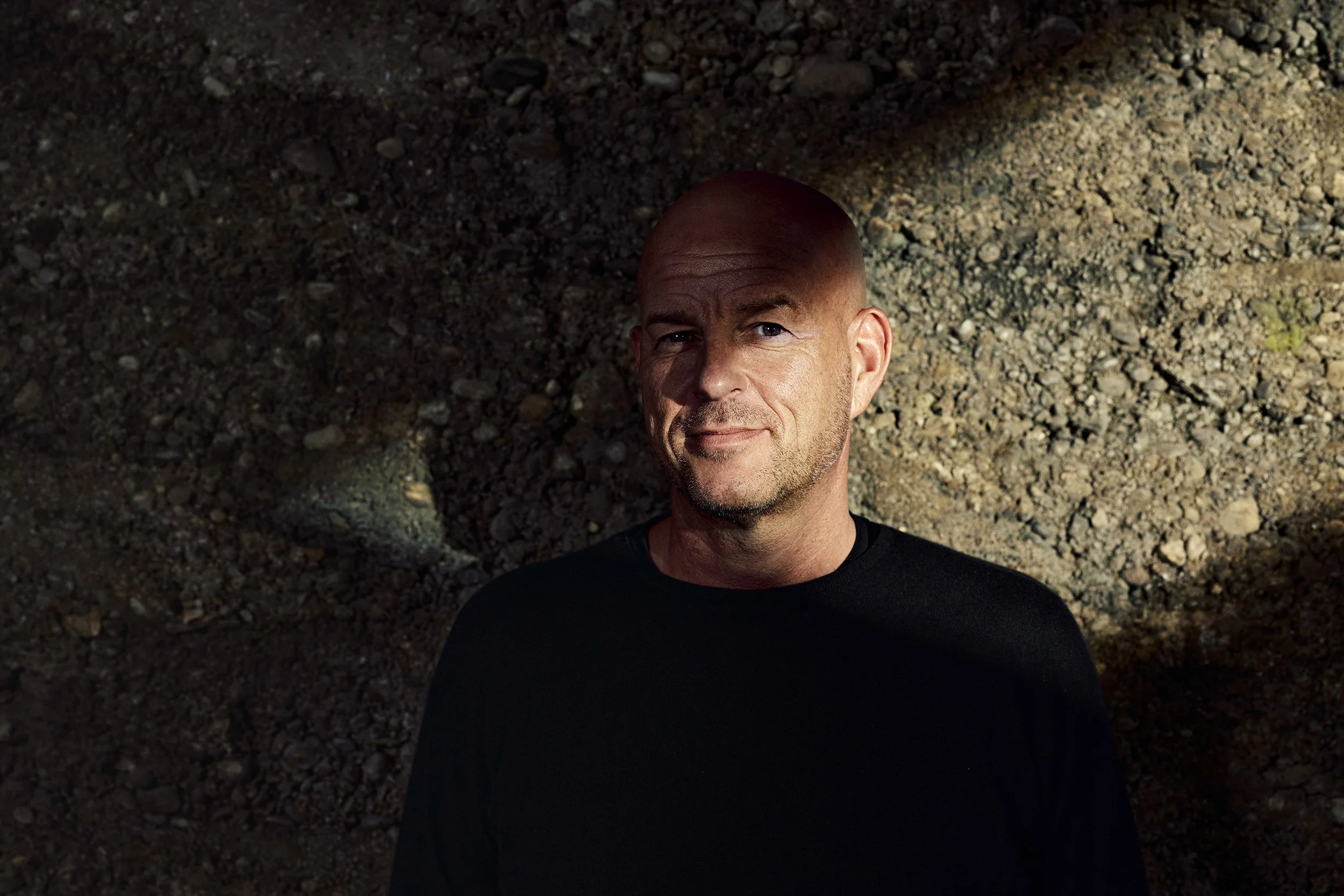 Ein Mann mit Glatze trägt ein schwarzes Shirt und steht vor einem dunklen, erdigen Hintergrund.