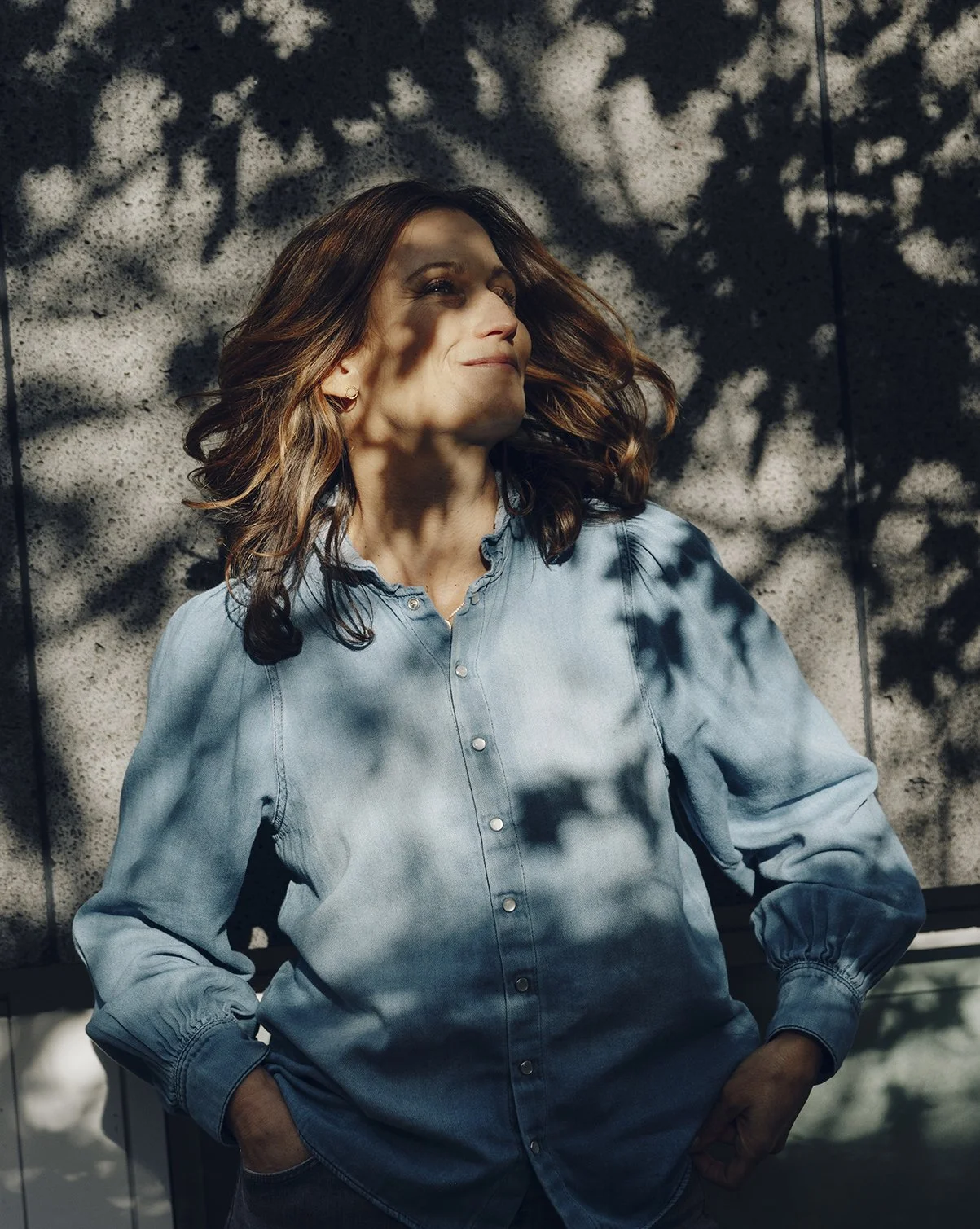 Frau steht im Schatten eines Baumes, Sonnenlicht wirft Schatten auf ihr Gesicht, sie trägt eine blaugeblendete Hemdbluse, lächelt leicht und wirkt entspannt.