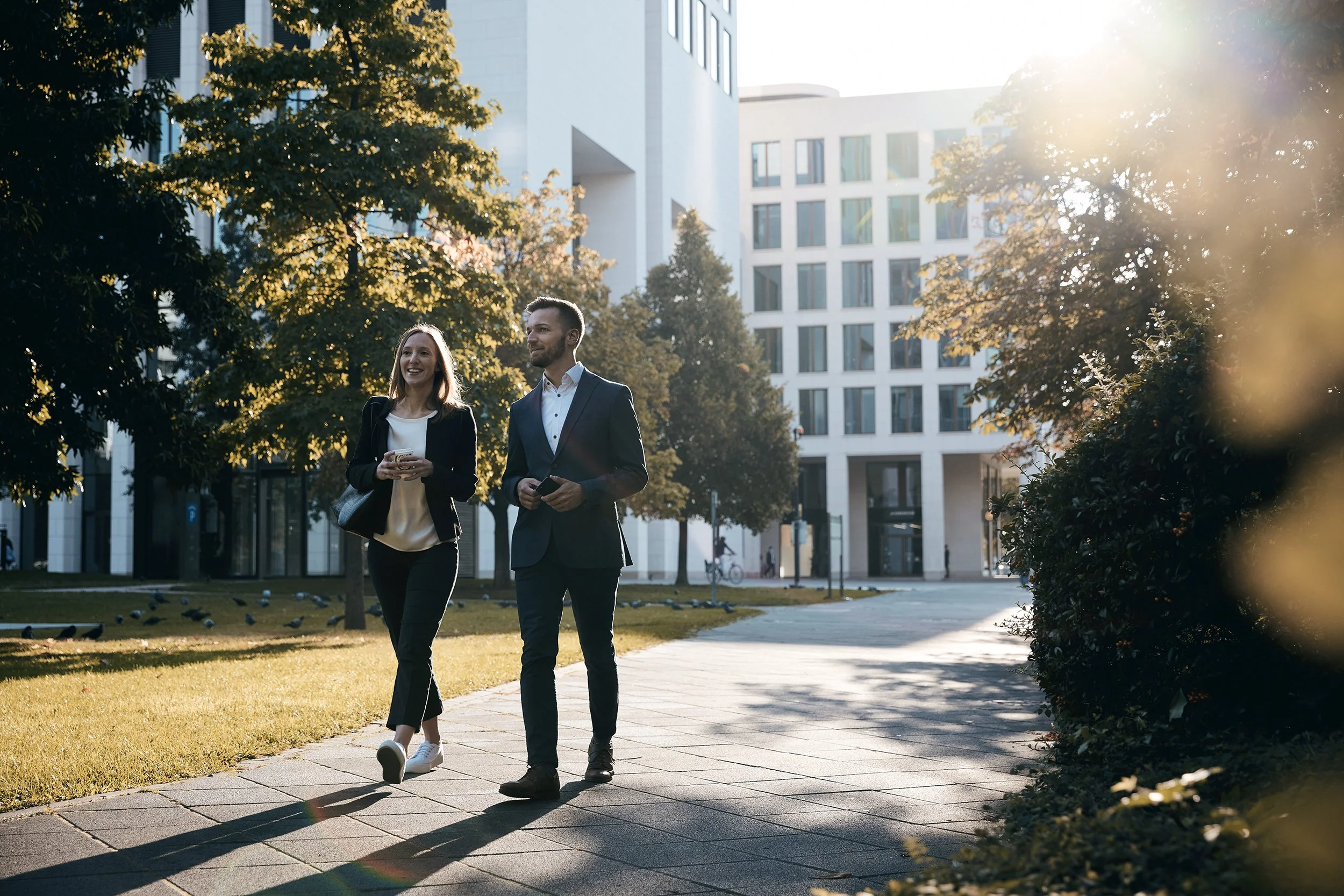 Zwei berufstätige Menschen, eine Frau und ein Mann, gehen auf einem Gehweg in einer modernen Stadtumgebung bei Sonnenuntergang, lächeln und unterhalten sich.