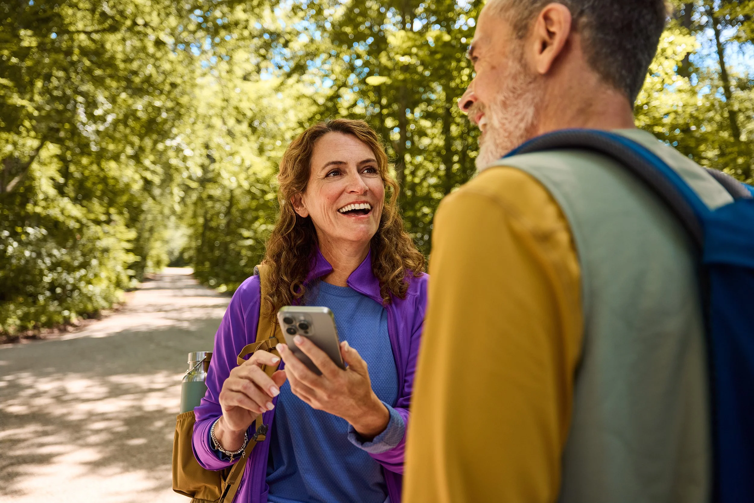 Zwei wandernde Personen unterhalten sich auf einem Waldweg, die Frau zeigt auf ihr Smartphone, beide tragen Wanderkleidung und Rucksäcke.
