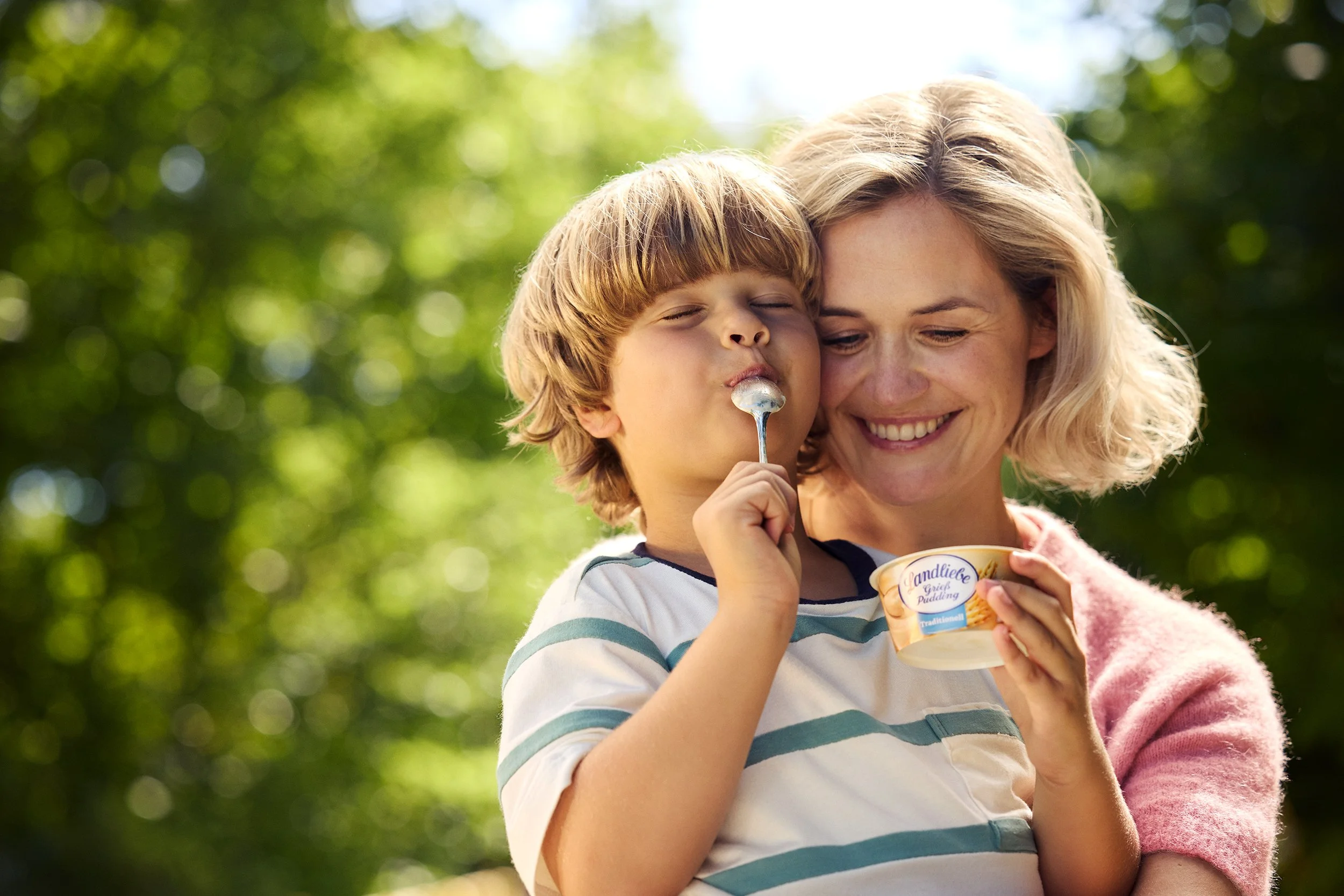 Eine lachende Frau umarmt einen Jungen, der Eiscreme isst, im Freien bei Sonnenlicht, mit grünem Bokeh-Hintergrund.