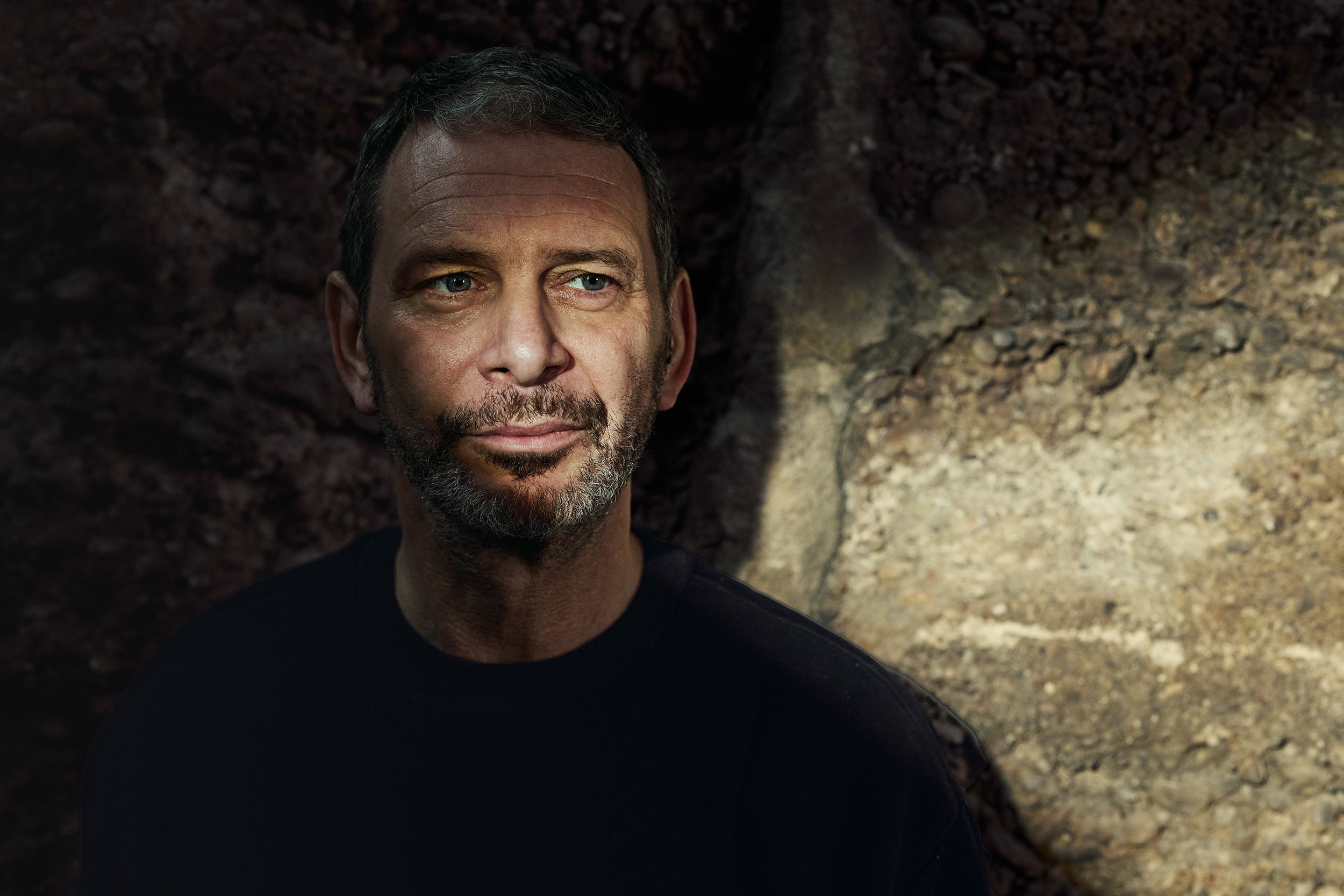 Porträt eines Mannes mit dunklem Haar und Bart vor einem felsigen Hintergrund, blickt nach rechts.
