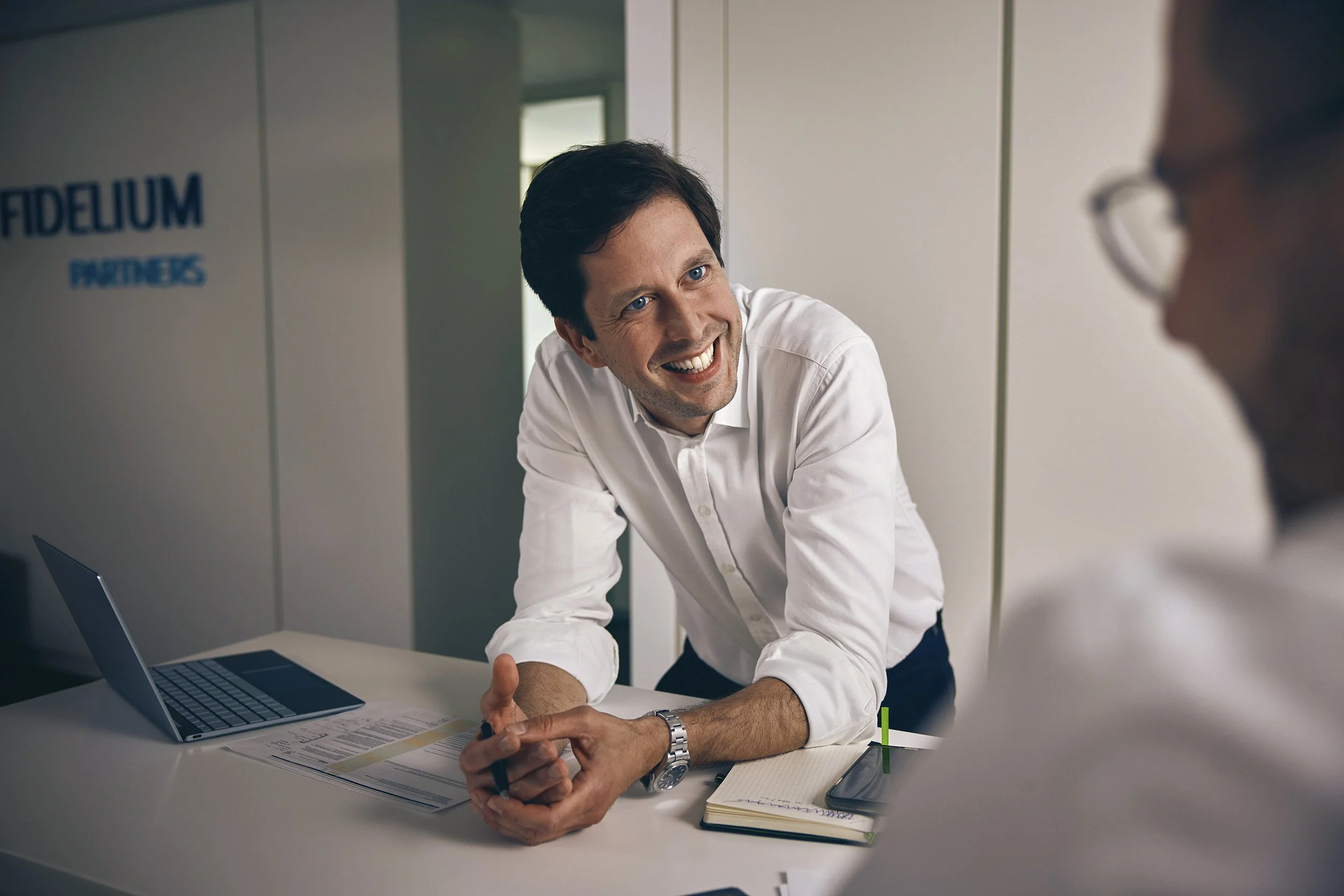 Zwei Männer unterhalten sich in einem Büro. Der Mann auf der linken Seite lächelt, trägt ein weißes Hemd und hat dunkle Haare. Der andere Mann sitzt rechts, trägt Brille, und seine Gesichtsausdruck ist nicht sichtbar. Es liegen ein Laptop, Notizbuch,