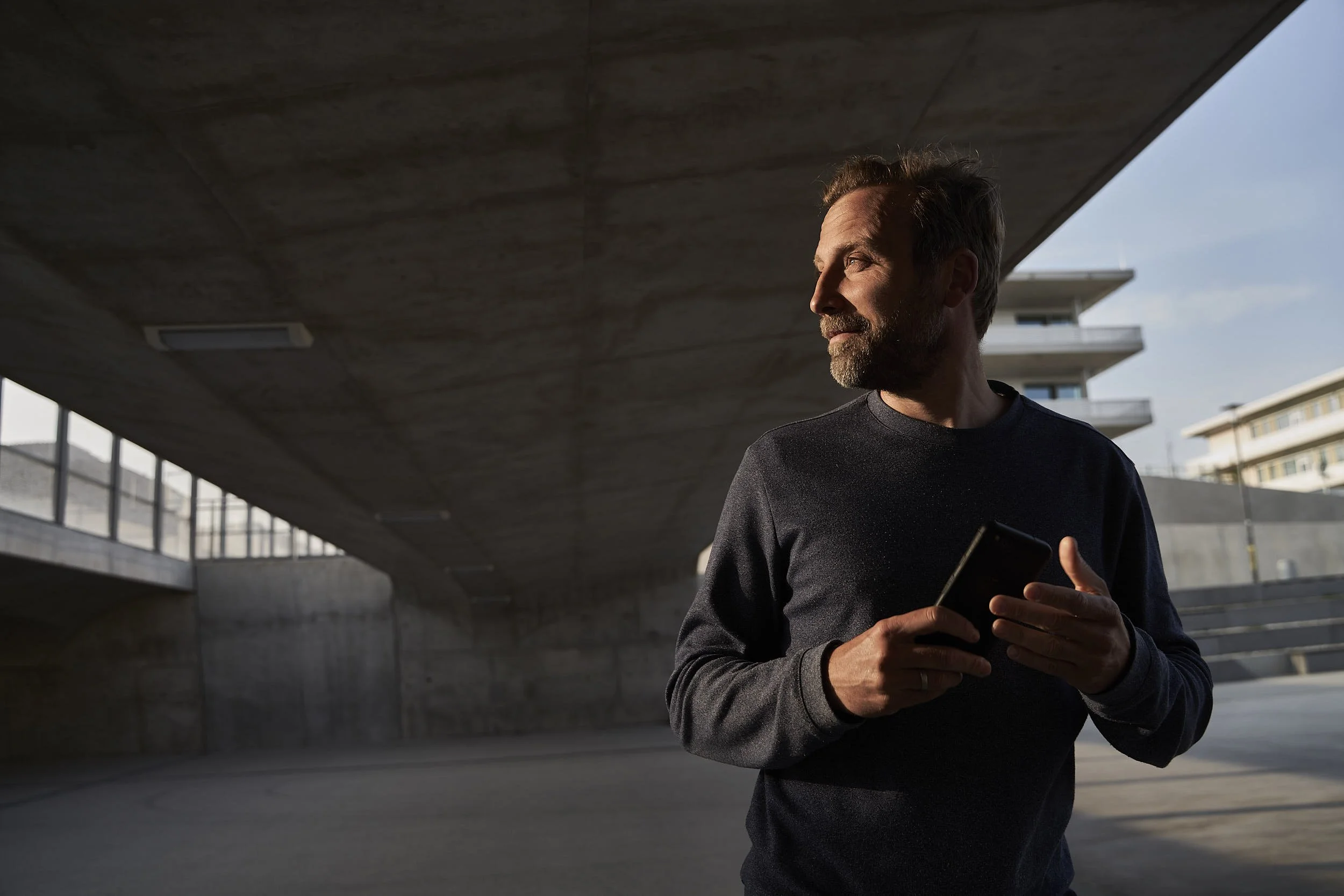Ein Mann mit grauem Bart und dunklem Pullover hält ein Smartphone und steht unter einer Überführung bei Sonnenlicht, im Hintergrund moderne Gebäude.