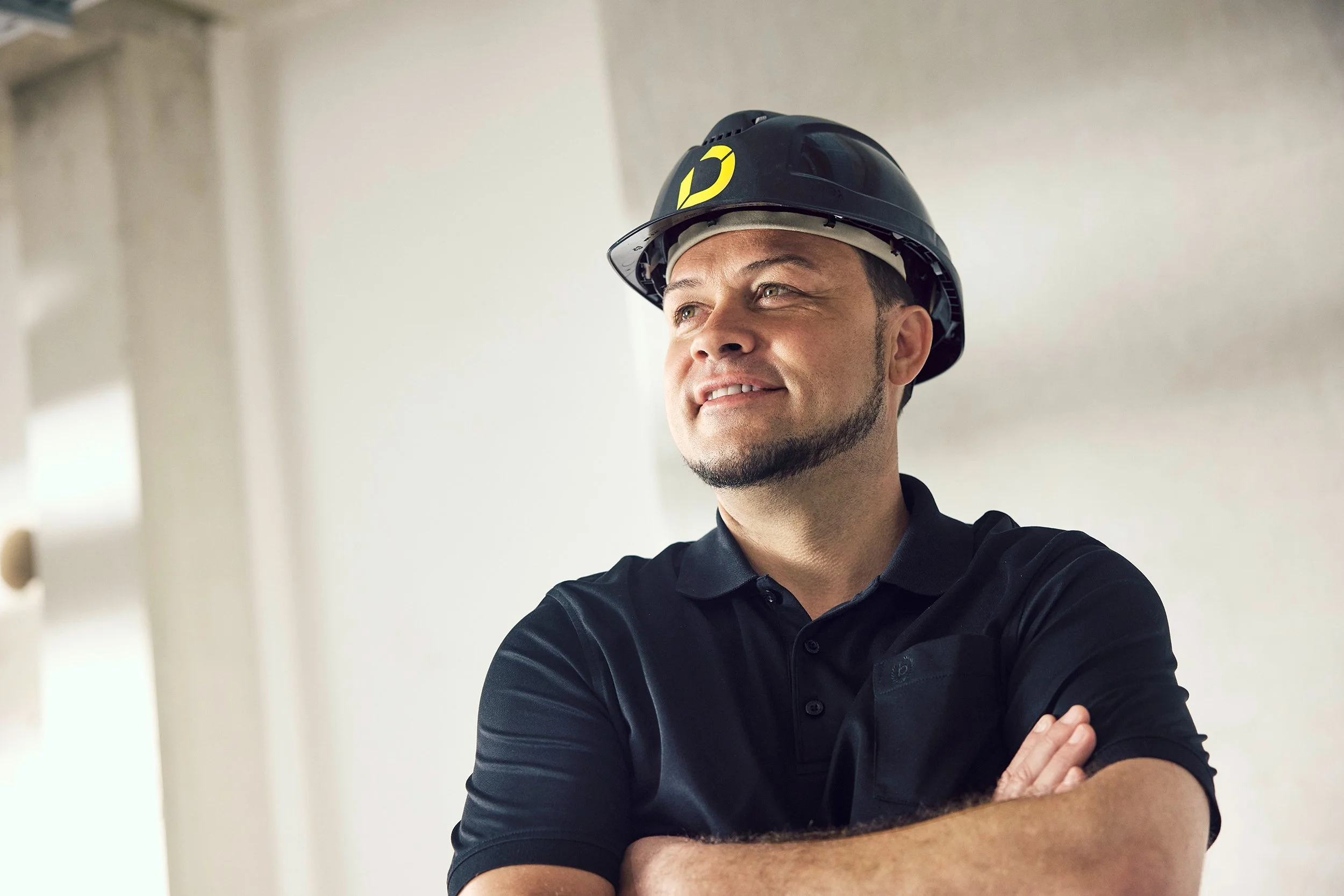 Mann trägt Schutzhelm und schwarze Polo-Hemd, steht mit verschränkten Armen und blickt zufrieden in die Ferne.