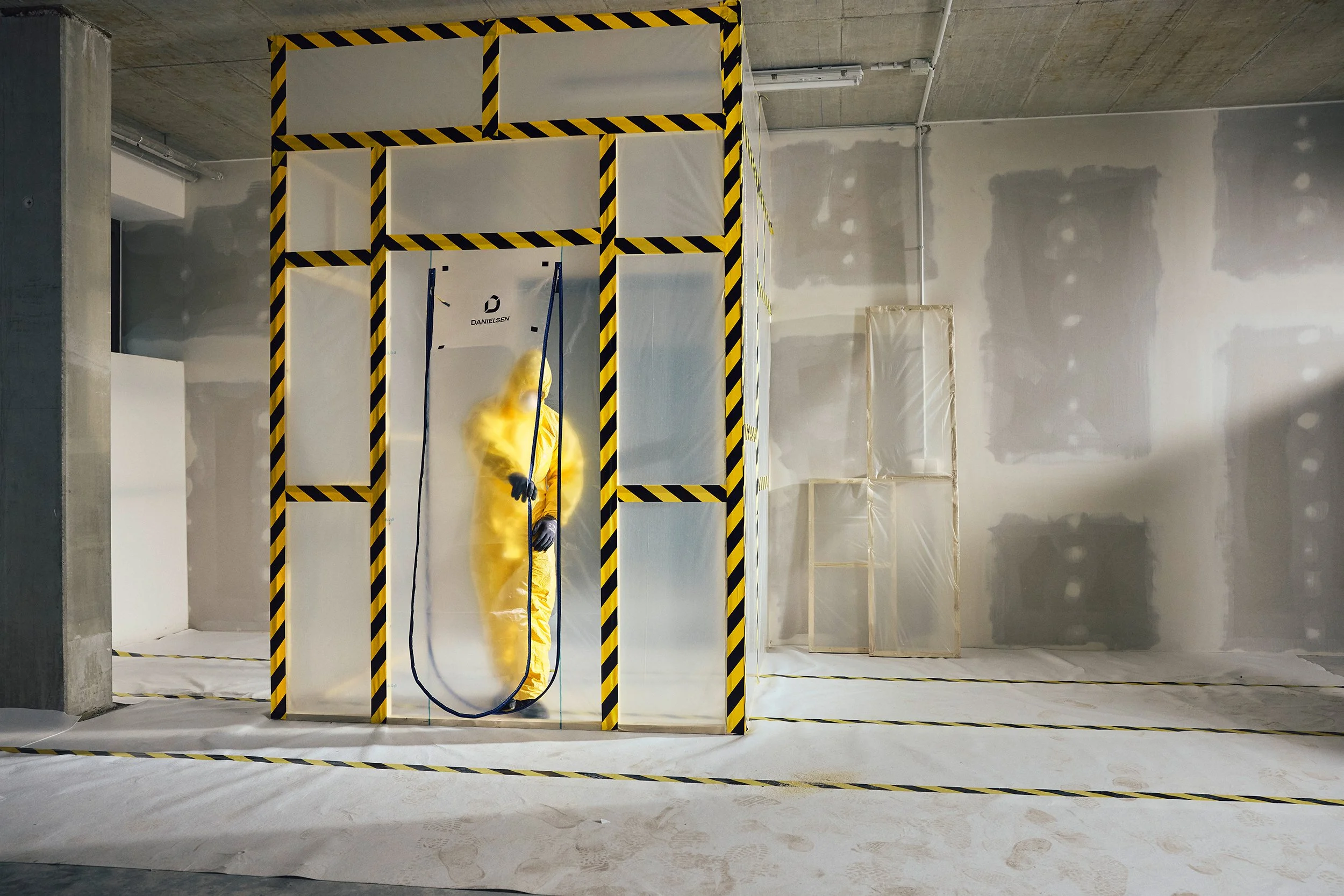 Ein Arbeiter in gelber Schutzkleidung steht in einem Raum und sprüht eine Substanz. Der Raum ist für die Bau- oder Renovierungsarbeiten vorbereitet, mit Abdeckungen auf dem Boden und weißen Platten an den Wänden.