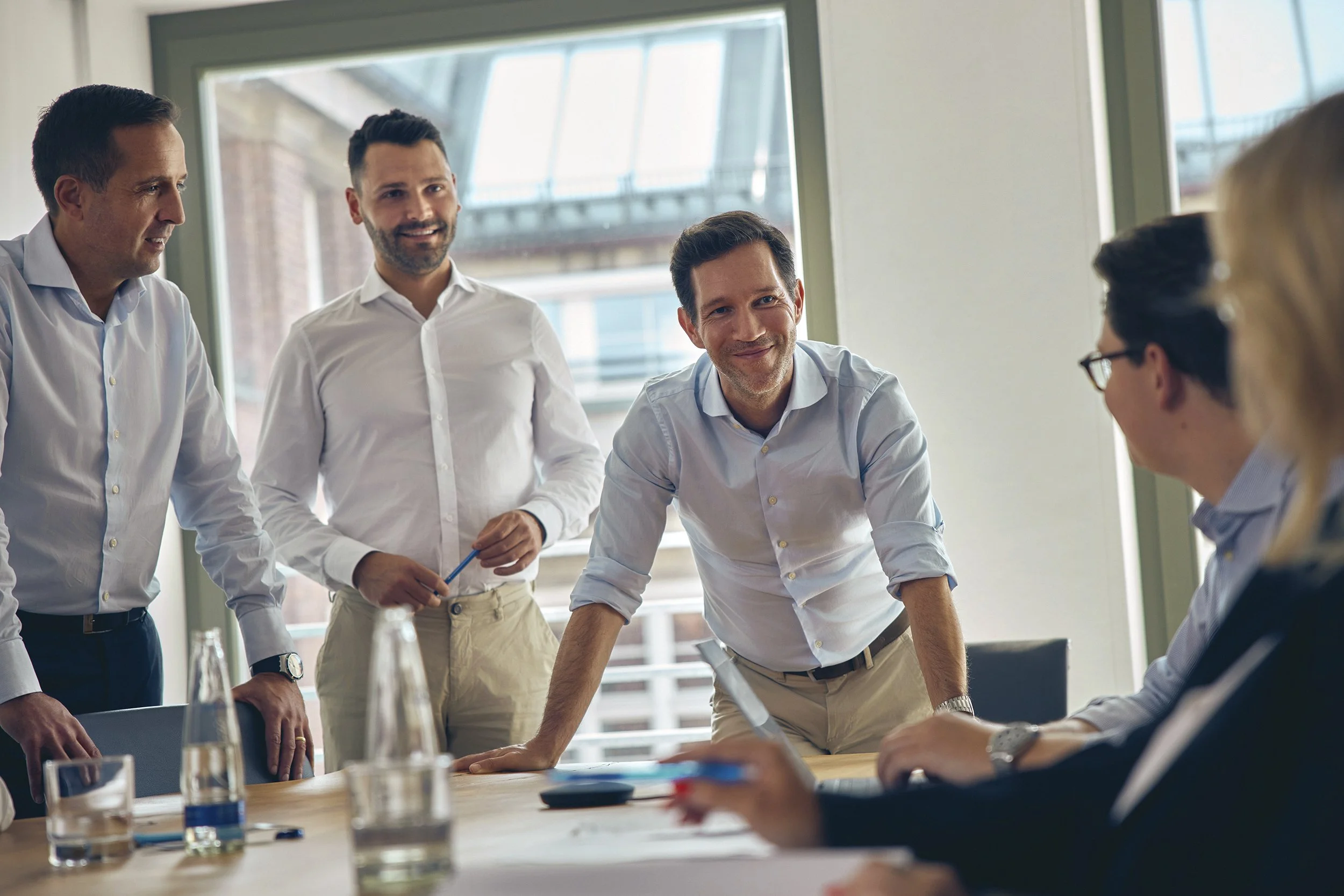 Vier Männer in einem Büro diskutieren bei einem Meeting, zwei sitzen an einem Tisch, einer steht und lehnt sich darauf, während der andere mit einem Laptop arbeitet.