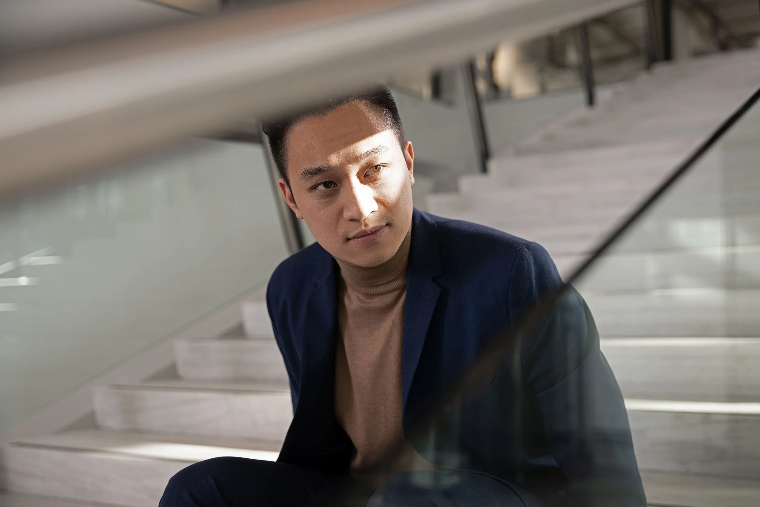 Ein junger Mann in dunklem Anzug und hellbraunem Rollkragenpullover sitzt auf einer Treppe, blickt nach oben und hat einen nachdenklichen Ausdruck.