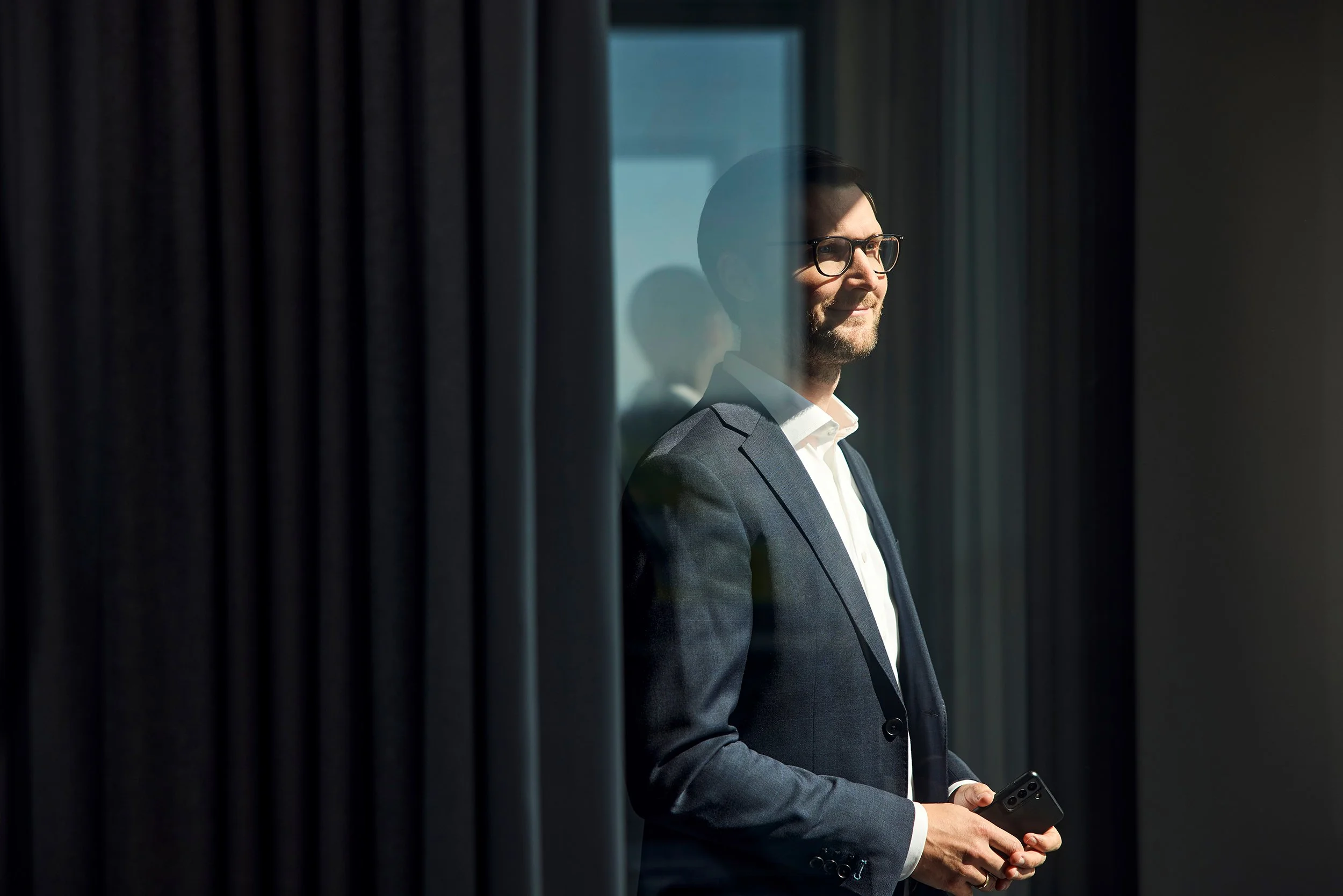 Mann in Anzug mit Brille und Smartphone, blickt durch eine Scheibe, vor dunklen Vorhängen.