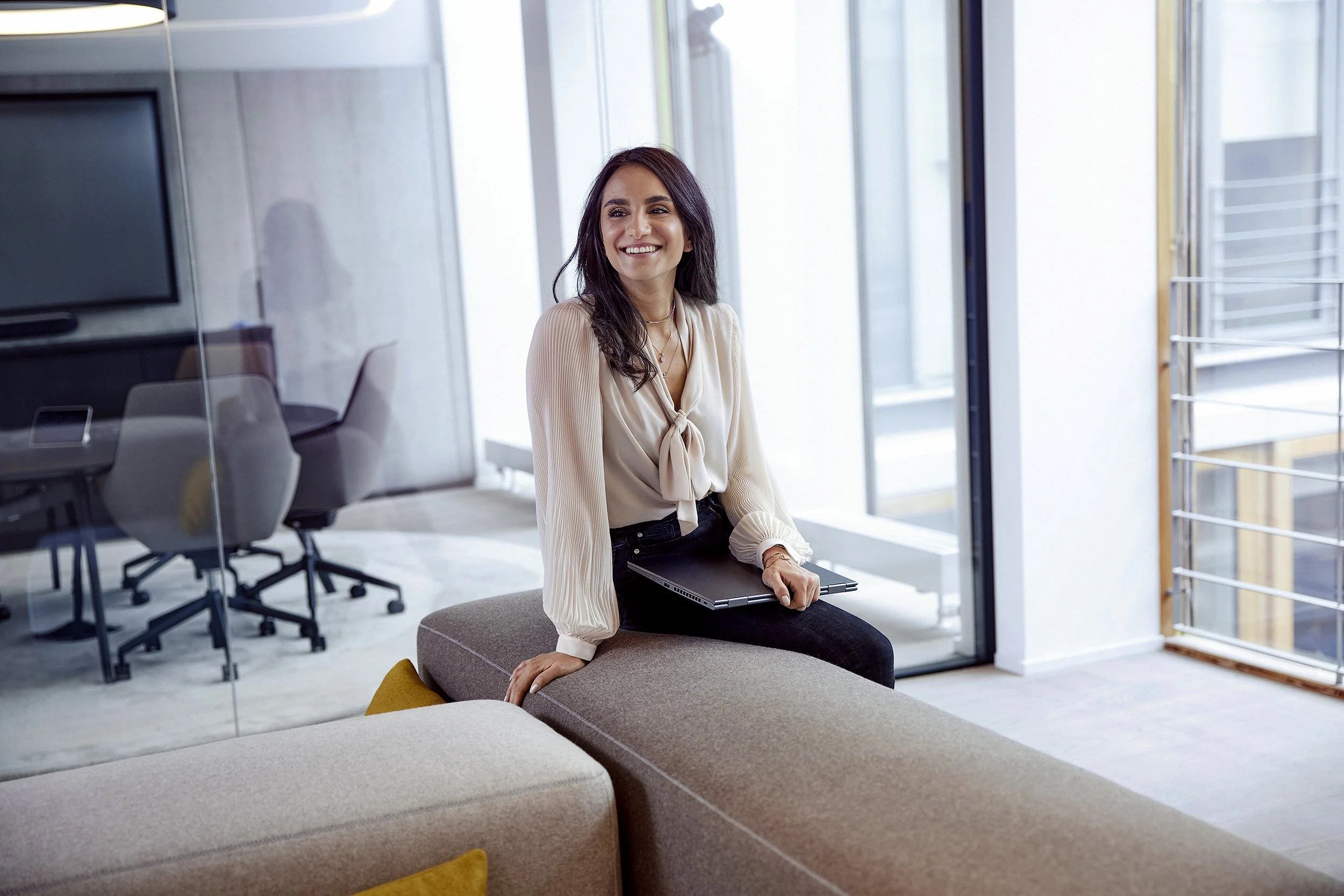 Eine lächelnde Frau sitzt in einem modernen Büro, trägt eine cremefarbene Bluse und hält einen Laptop auf ihrem Schoß. Im Hintergrund sind Büroarbeitsplätze und große Fenster