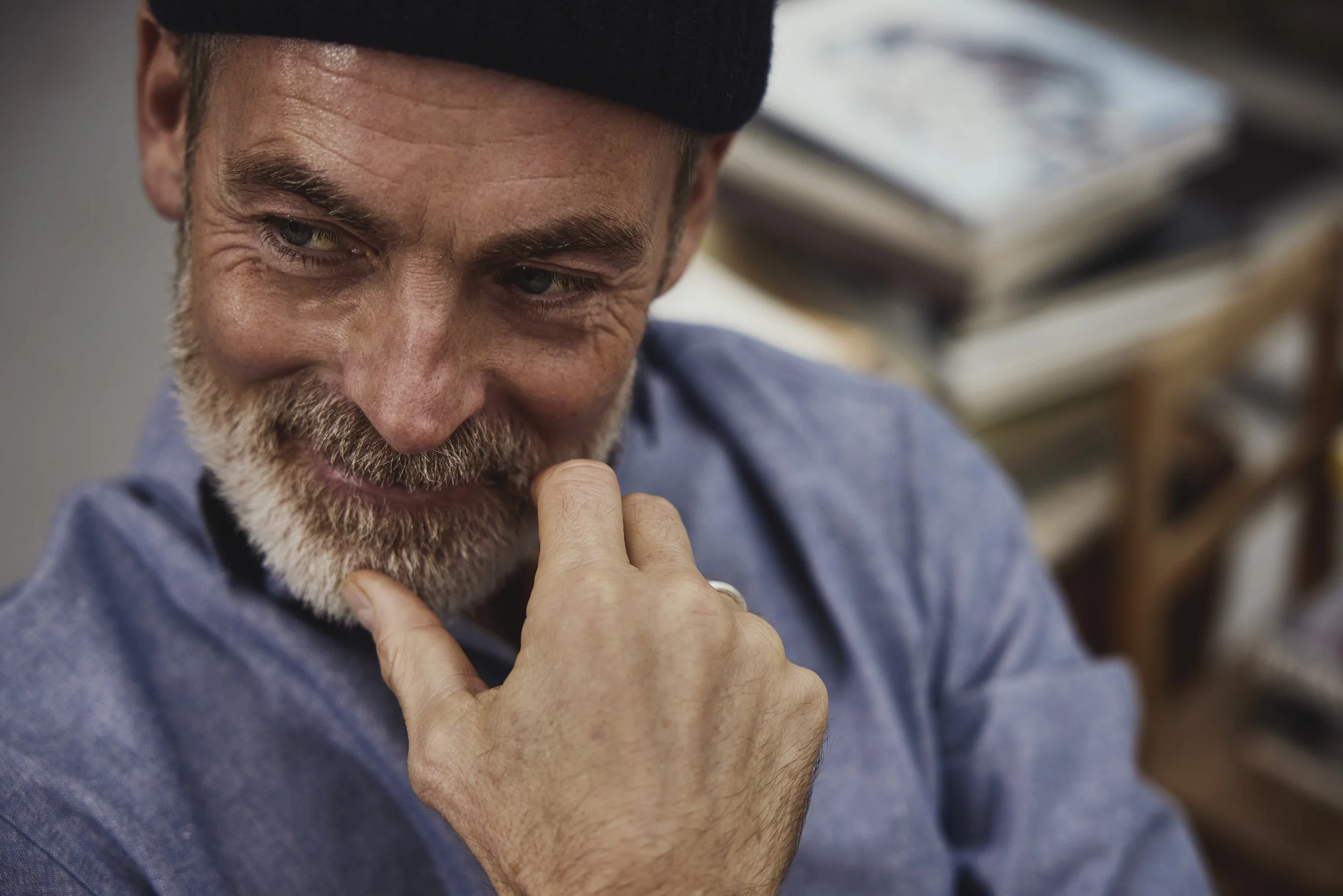 Nahaufnahme eines älteren Mannes mit grauem Bart, der an einem Kinn nachdenklich die Hand hält, während er nach unten blickt, im Hintergrund sind Bücher in Regalen.