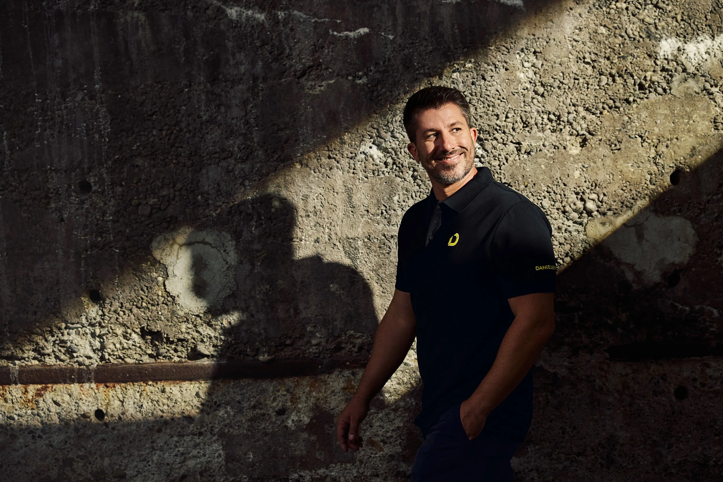 Ein lachender Mann mit dunklem Polo-Shirt vor einer sandigen Wand, Schatten wirft sich auf die Wand.