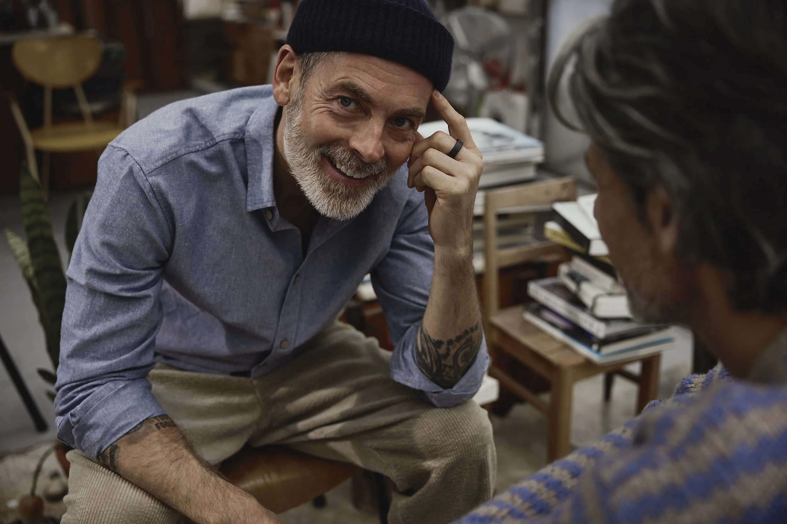 Zwei Männer im Gespräch, einer lächelt. Der Mann im Vordergrund trägt ein dunkles Hat, ein graues Hemd und beige Hosen, hat Tattoos am Arm und einen Bart.