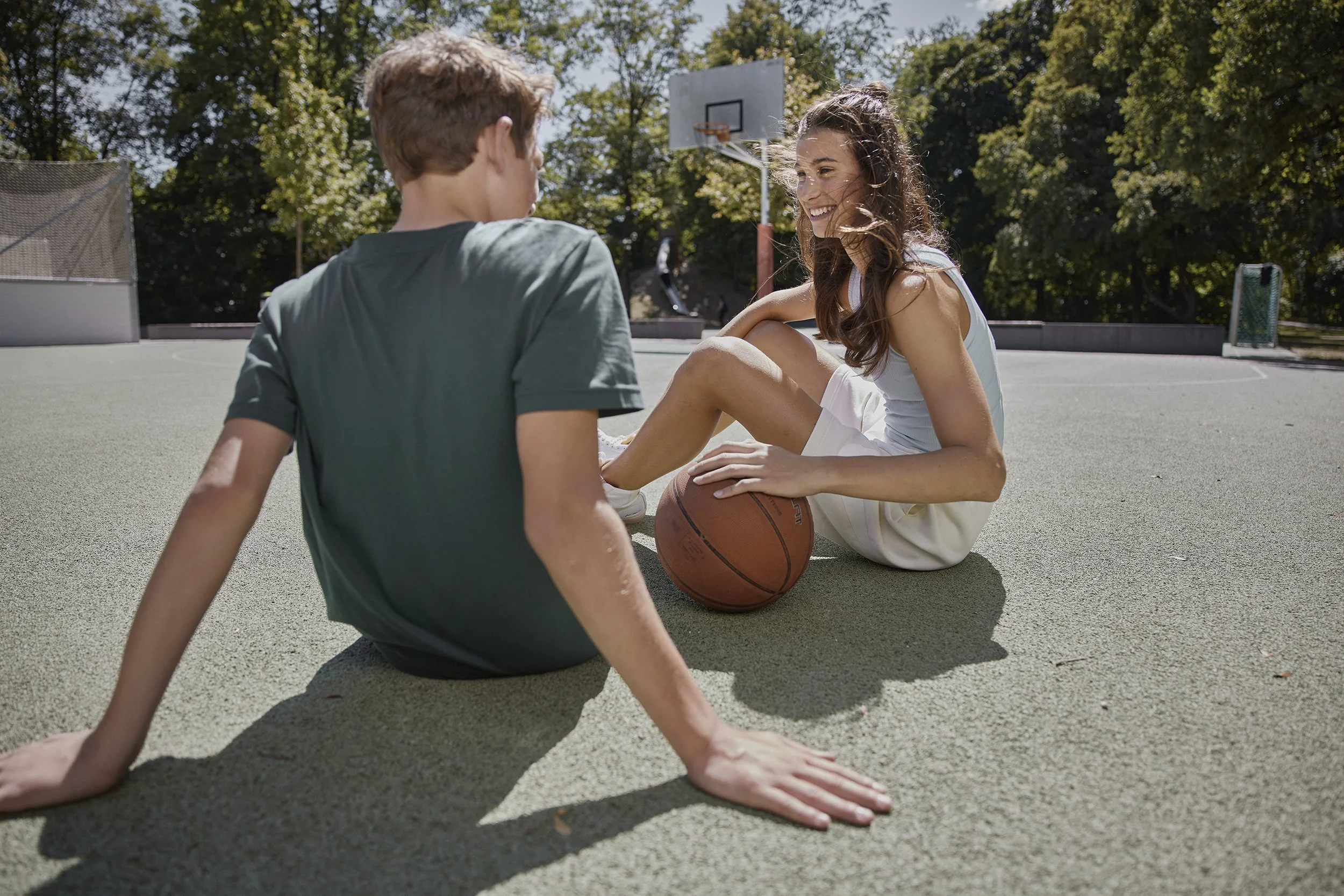 Ein Junge und ein Mädchen sitzen auf einem Basketballplatz, lachen und sprechen. Das Mädchen hat einen Basketball zwischen den Beinen und trägt ein weißes Sportoutfit, der Junge trägt ein graues T-Shirt.