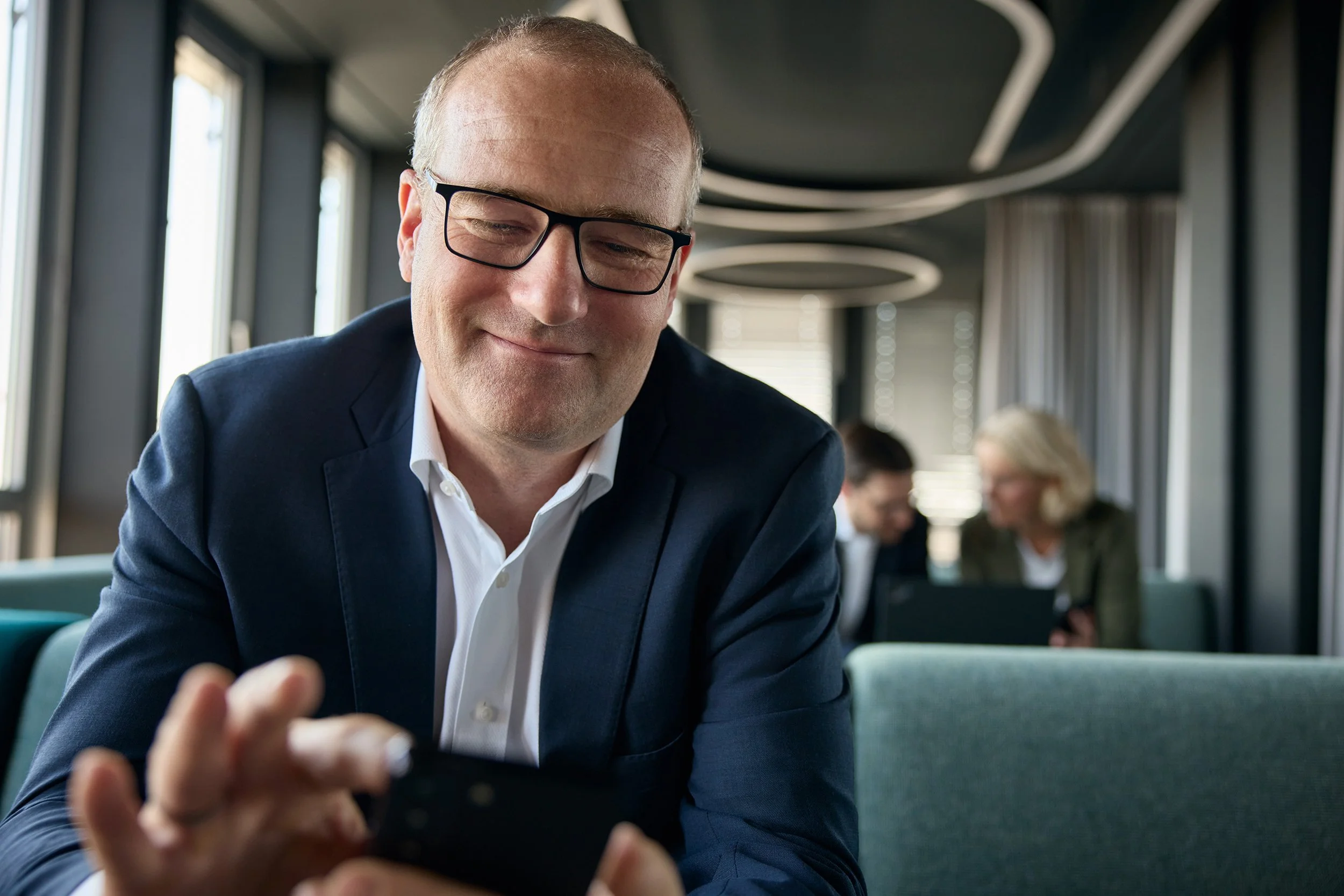 Ein Mann im Anzug und Brille schaut lächelnd auf sein Smartphone. Im Hintergrund sitzen zwei Personen an einem Tisch und schauen gemeinsam auf ein Laptop.