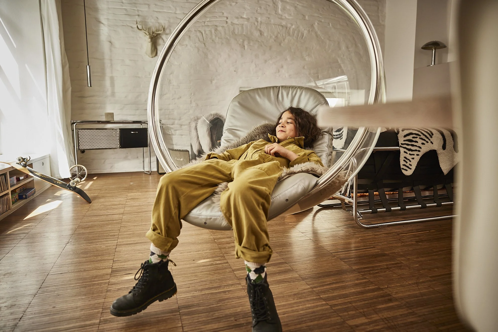 Ein Kind liegt entspannt in einer modernen, transparenten, hängenden Hängeliege in einem Wohnzimmer, mit Holzfußboden und weißen Wänden, in einer gemütlichen Atmosphäre.