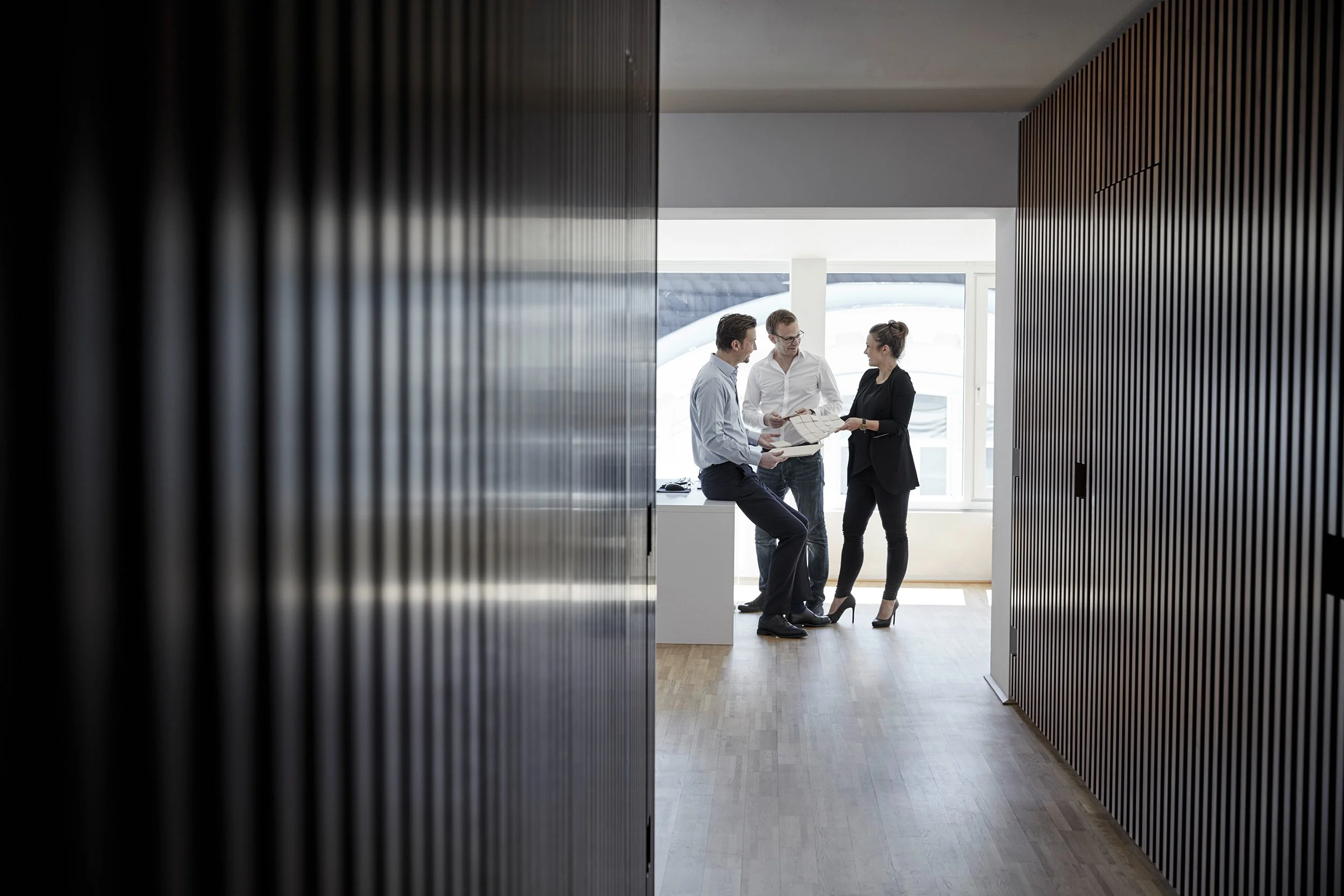 Drei Geschäftsleute diskutieren in einem modernen Büro mit Holzfußboden, Blick durch eine Türöffnung mit Holzverkleidung, bei Tageslicht.