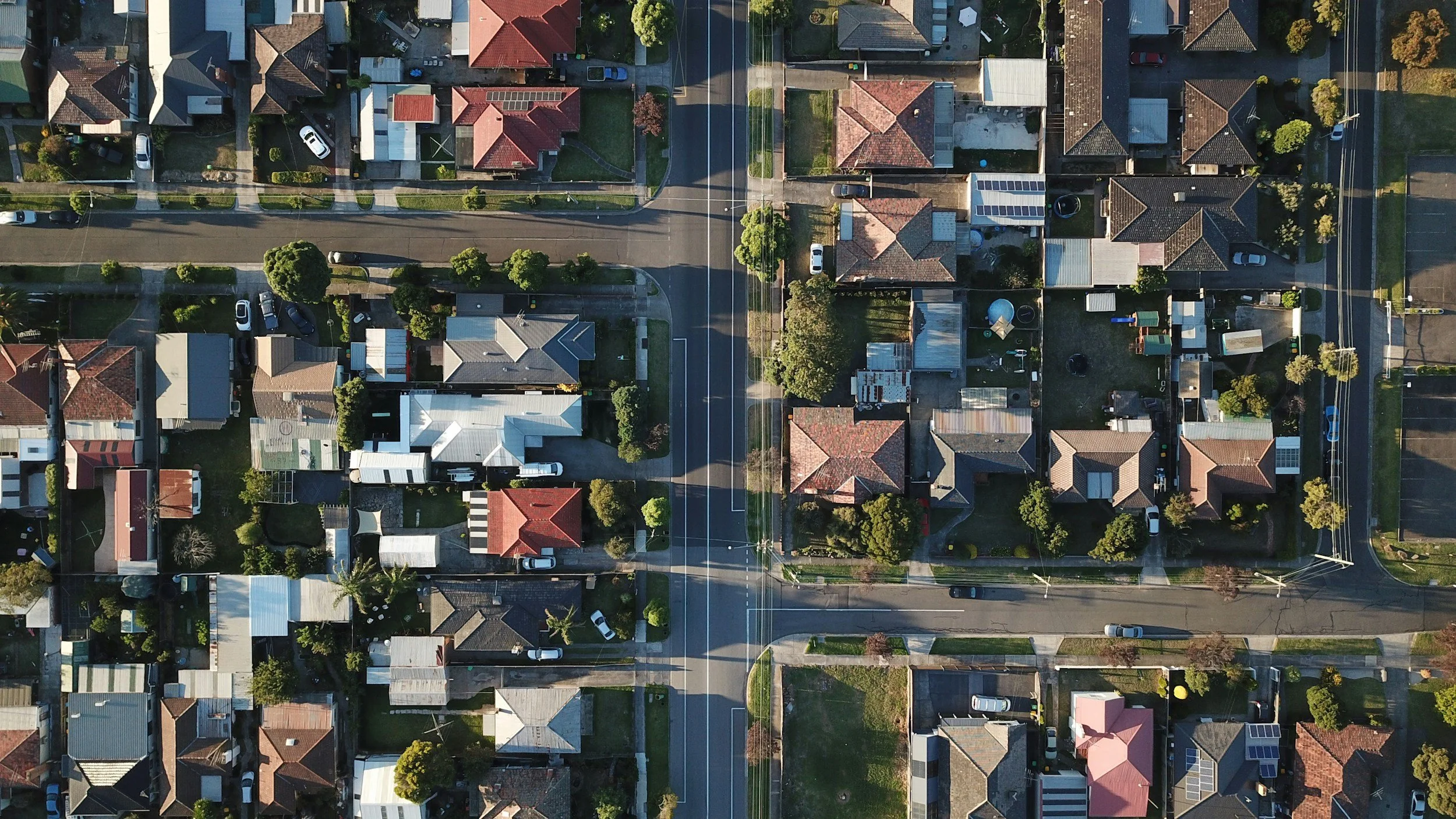 Aerial view of a suburban neighborhood showing multiple houses, roads, trees, and parked cars.