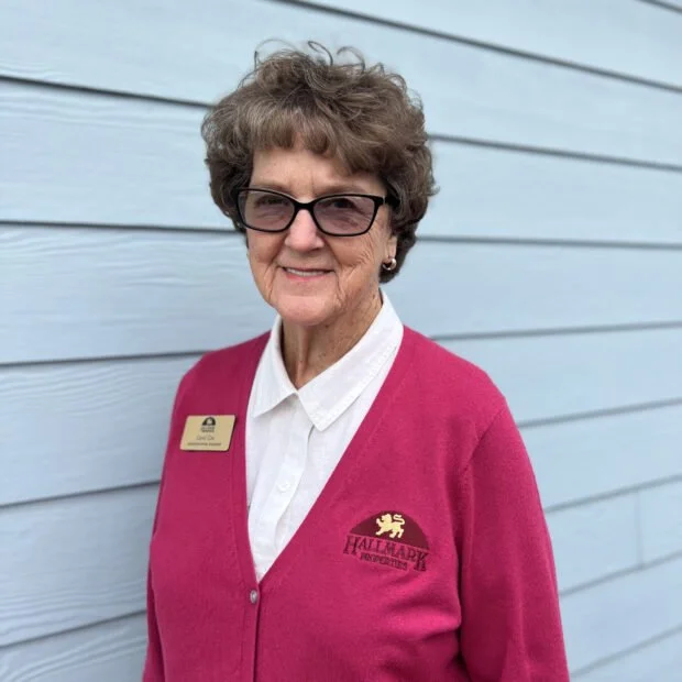 Older woman smiling, wearing glasses, a white blouse, and a pink cardigan with a Hallmark research logo and a name badge, standing in front of a light blue wooden wall.