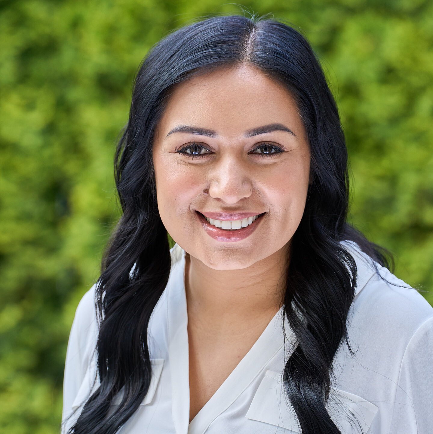 Smiling woman with long black hair in white shirt outdoors with green blurred background.