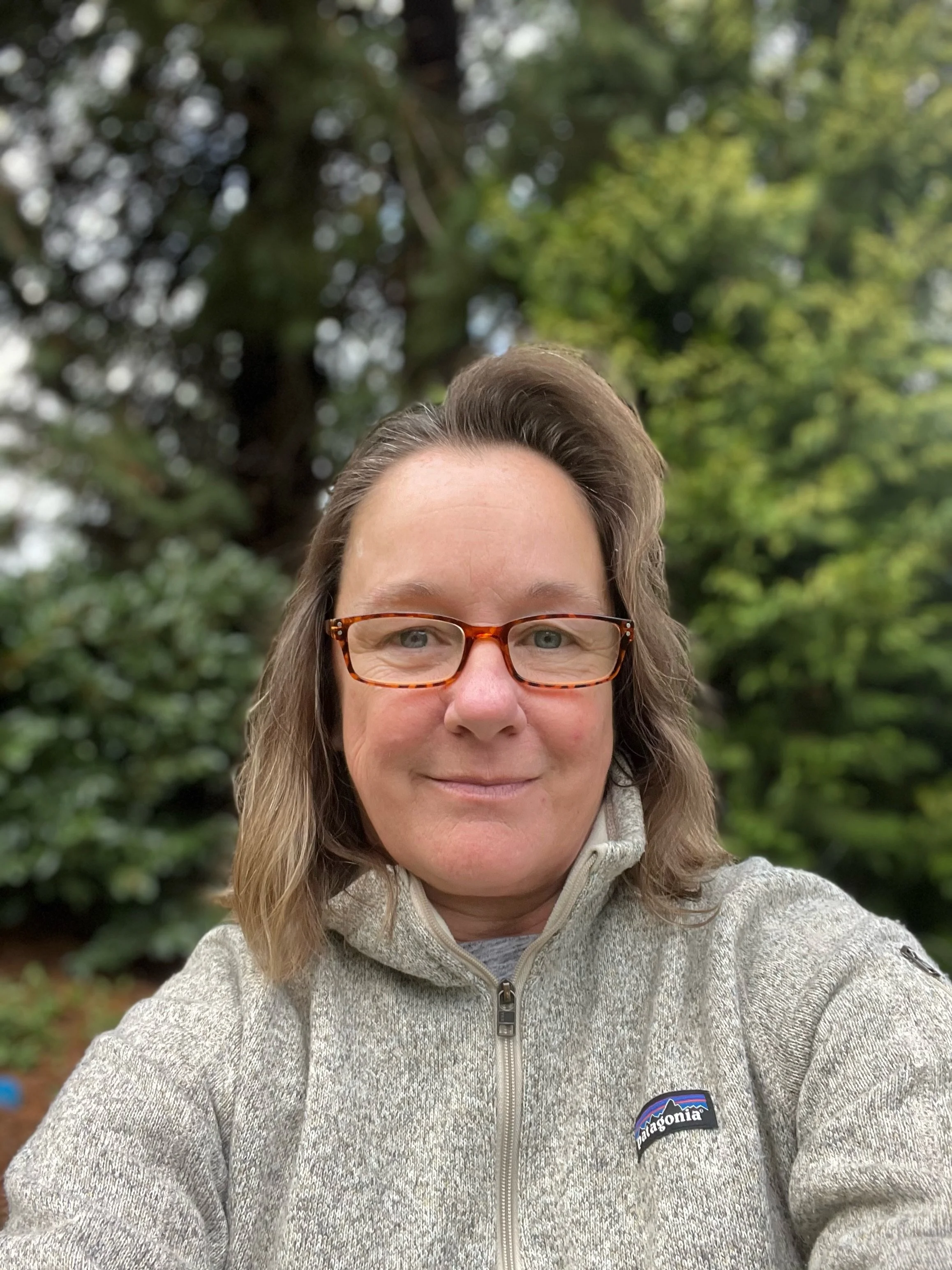 A woman with light brown hair and glasses taking a selfie outdoors in front of a large tree and green foliage. She is wearing a gray Patagonia fleece jacket.