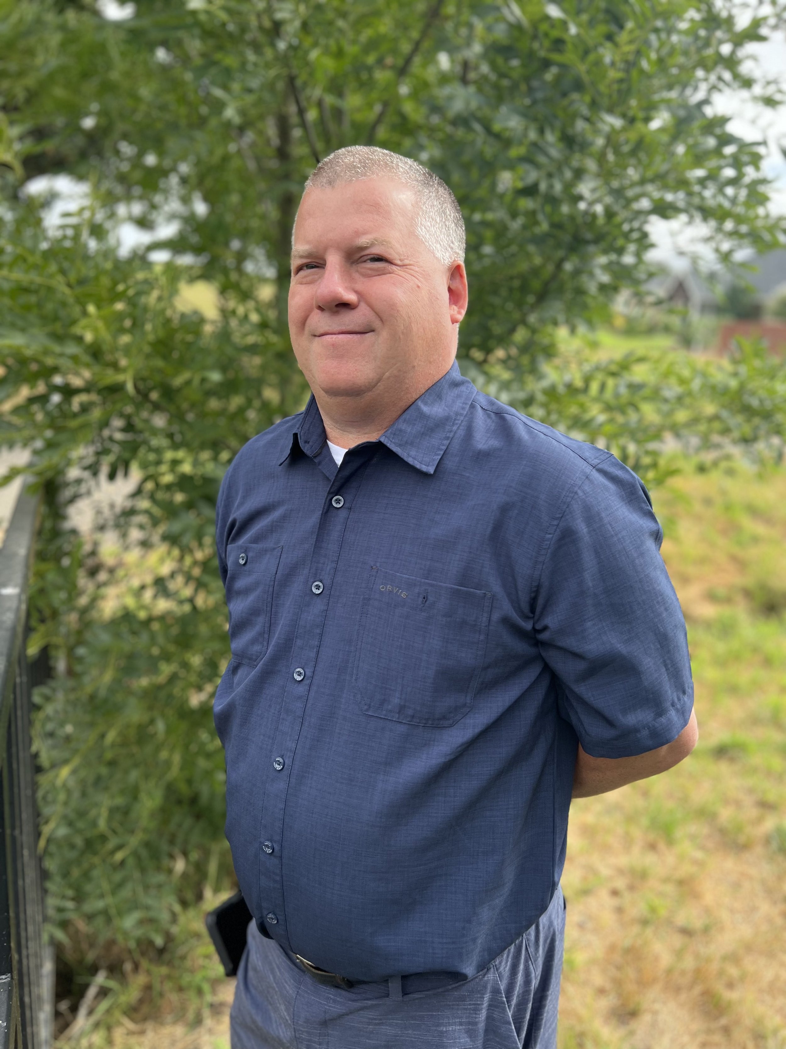 A man standing outdoors in a natural setting with green trees and foliage in the background, wearing a navy blue button-up shirt and patterned pants.