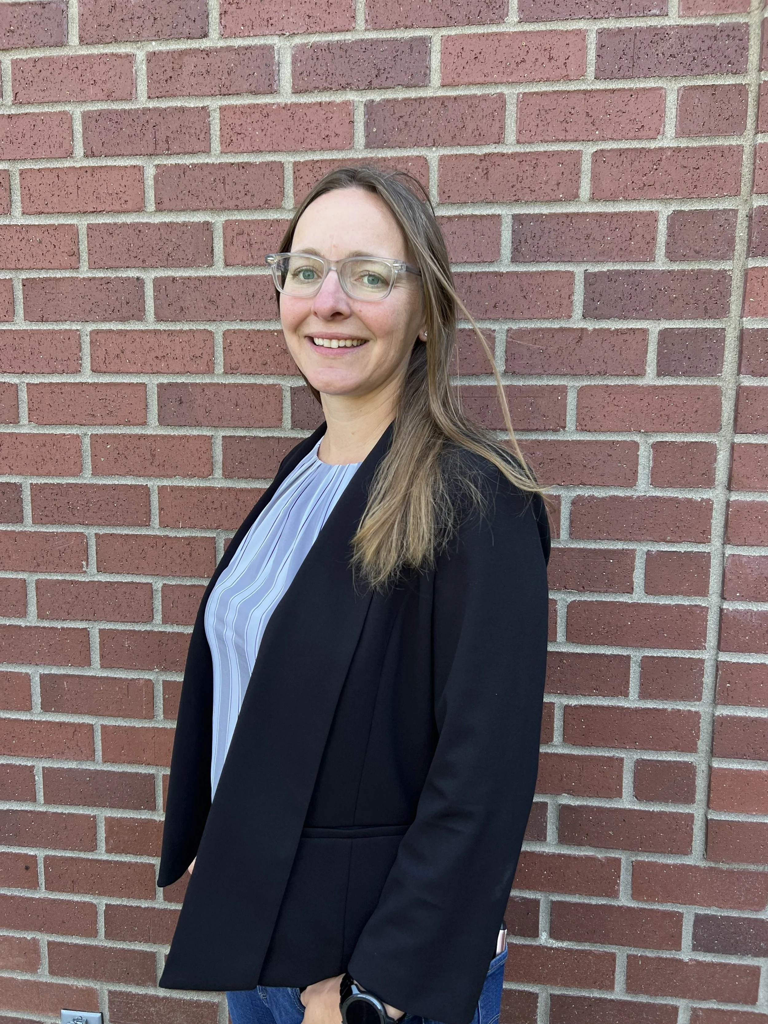 A woman with light skin, long light brown hair, wearing glasses, a black blazer, a blue and white striped shirt, and blue jeans, standing in front of a red brick wall, smiling at the camera.