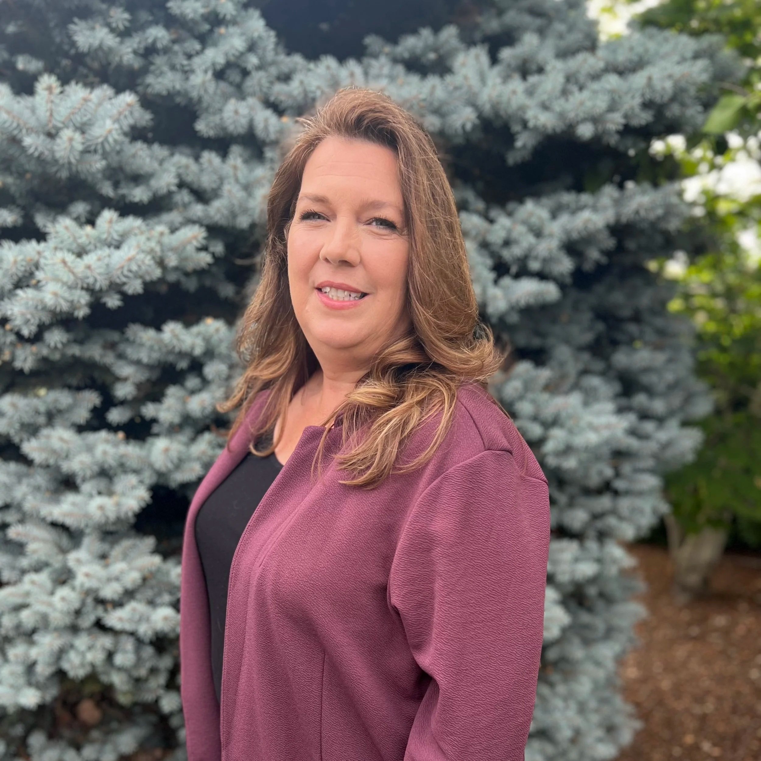 A woman with long, wavy hair smiling, wearing a pink blazer, stands outdoors in front of a large blue spruce tree and some green foliage.