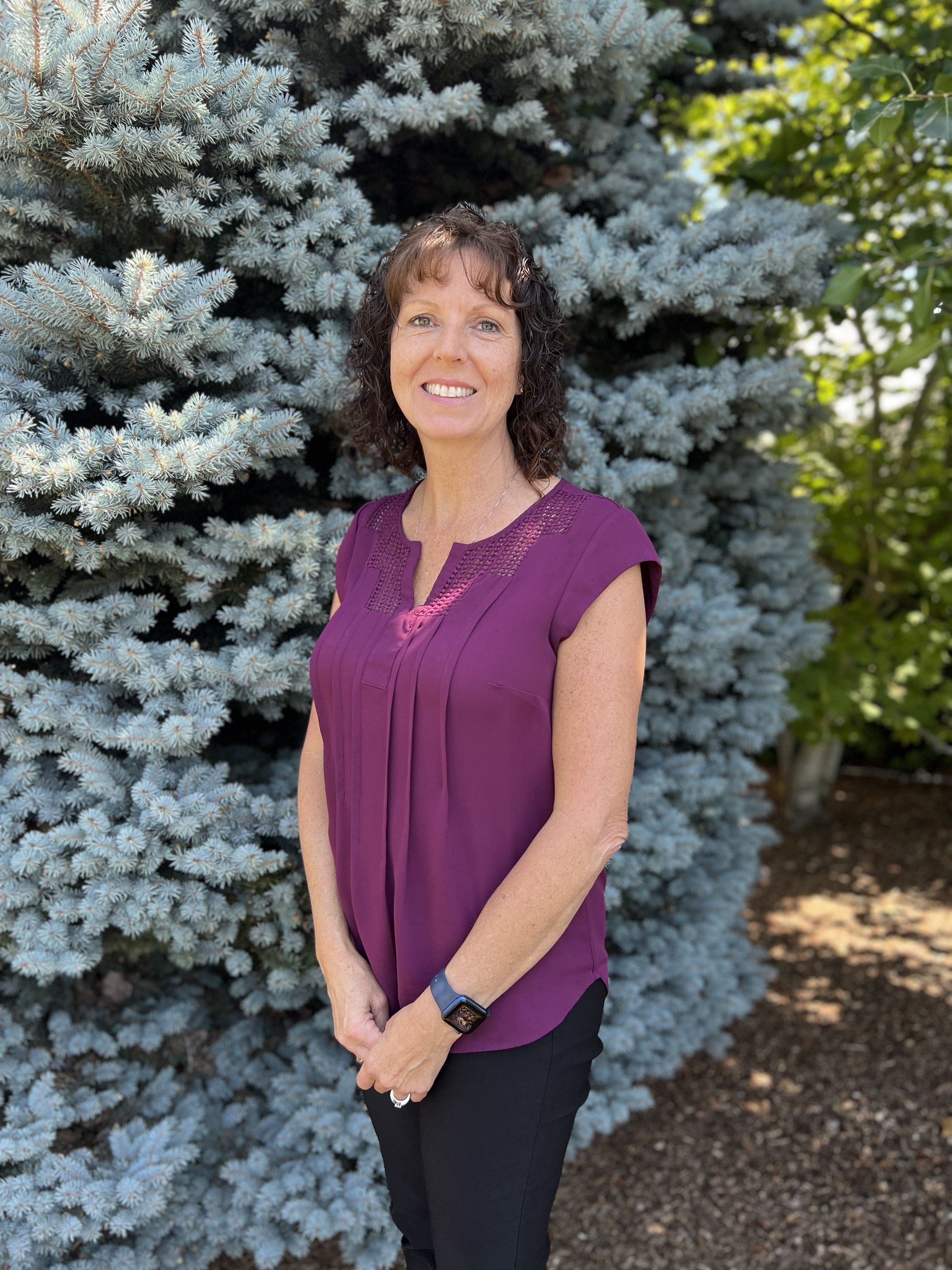 A woman with dark curly hair and light skin smiling outdoors, wearing a purple blouse and black pants, standing in front of a blue spruce tree with green leaves in the background.