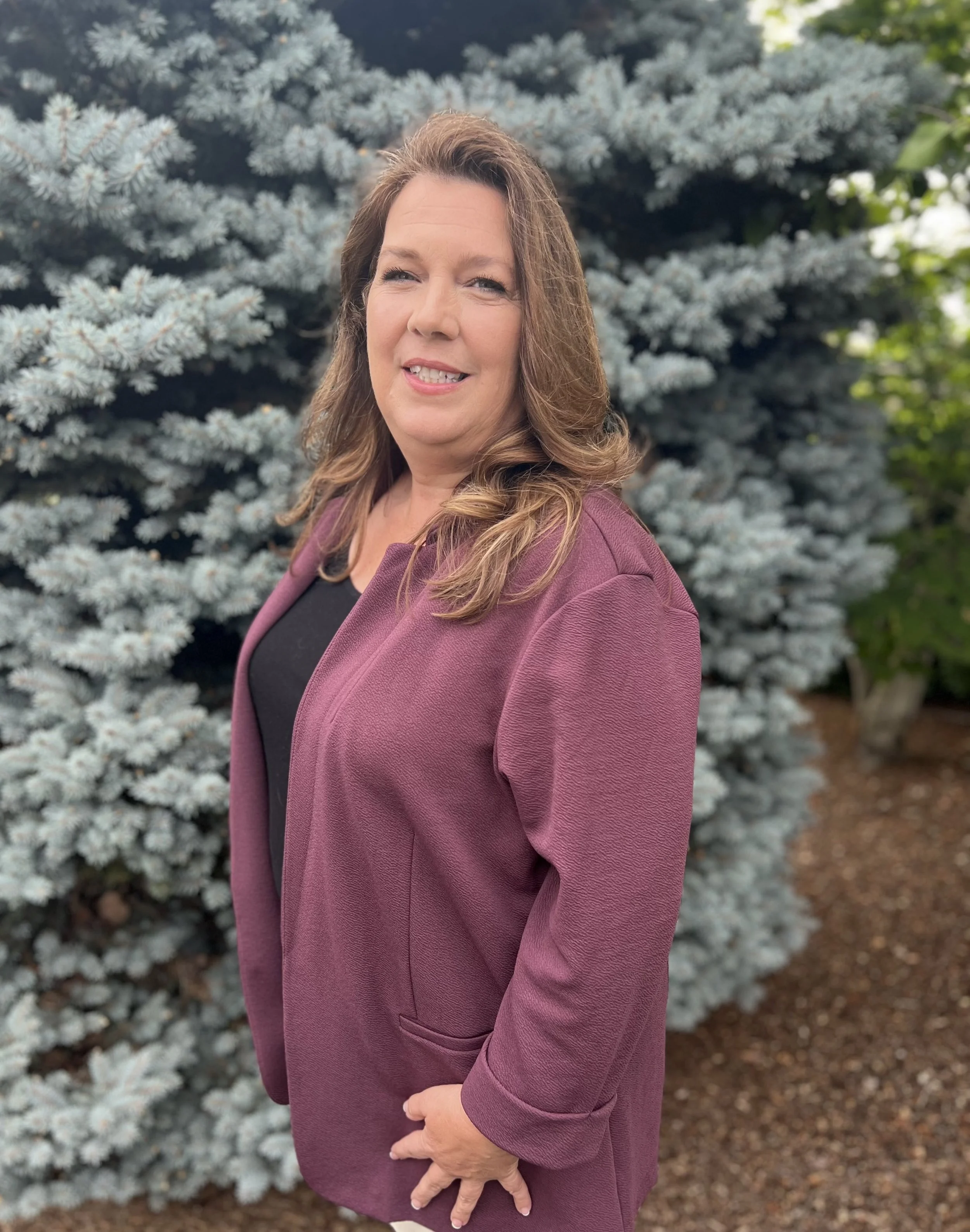 Portrait of a woman with wavy brown hair and blue eyes, wearing a black top and a purple jacket, standing outdoors in front of a blue spruce tree with dense, silver-blue needles.