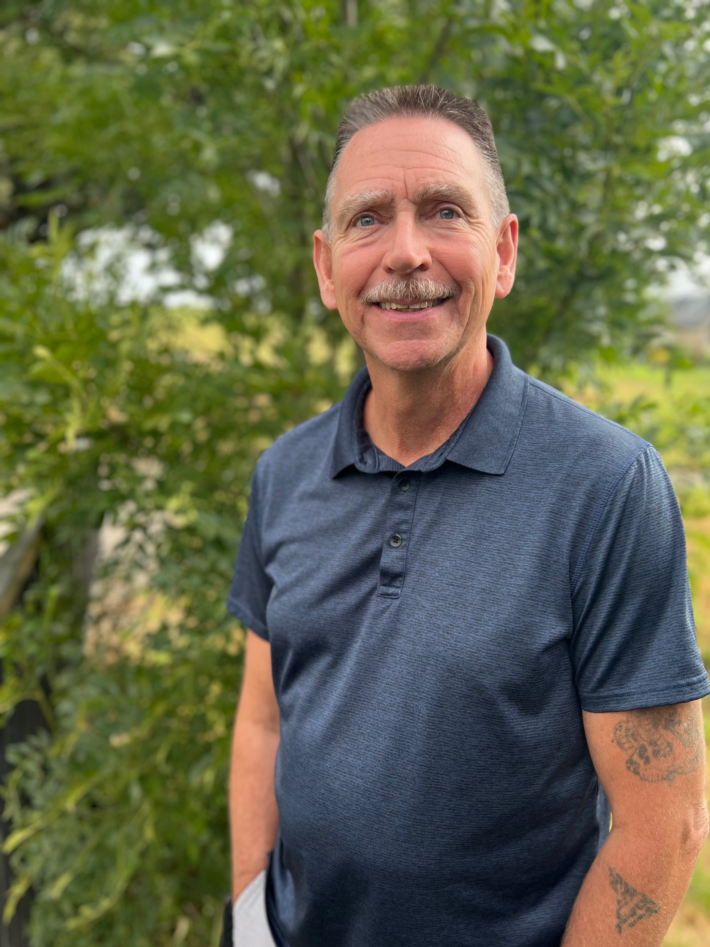 Close-up of a smiling middle-aged man with short dark hair, blue eyes, and a gray mustache, wearing a dark blue polo shirt, standing outdoors with green trees and foliage in the background.