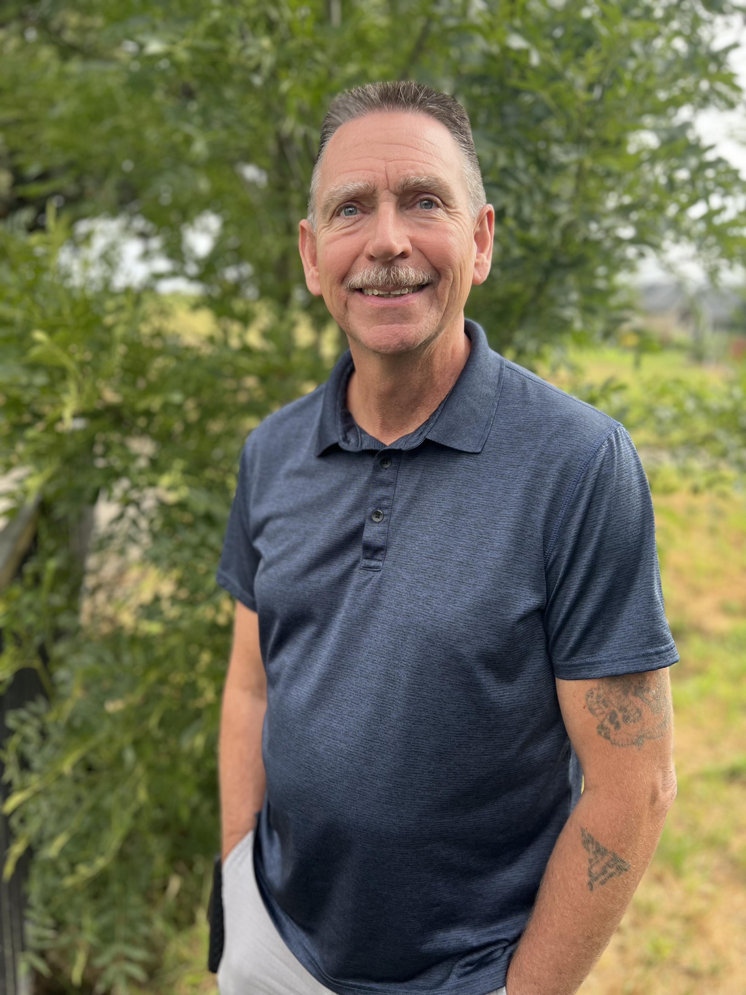 A smiling middle-aged man with short dark slicked-back hair, a mustache, and tattoos on his arm standing outdoors in front of green trees wearing a dark blue polo shirt.