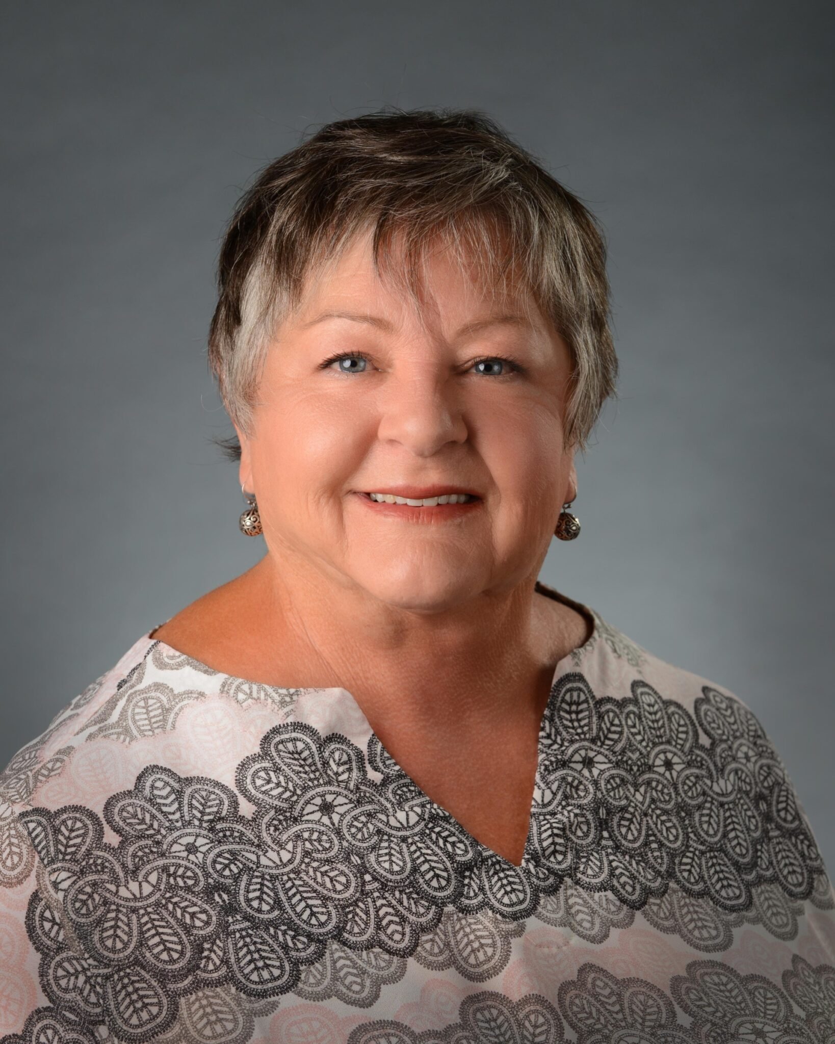 Close-up of a smiling woman with short gray hair wearing white and black patterned top and earrings against a gray background.