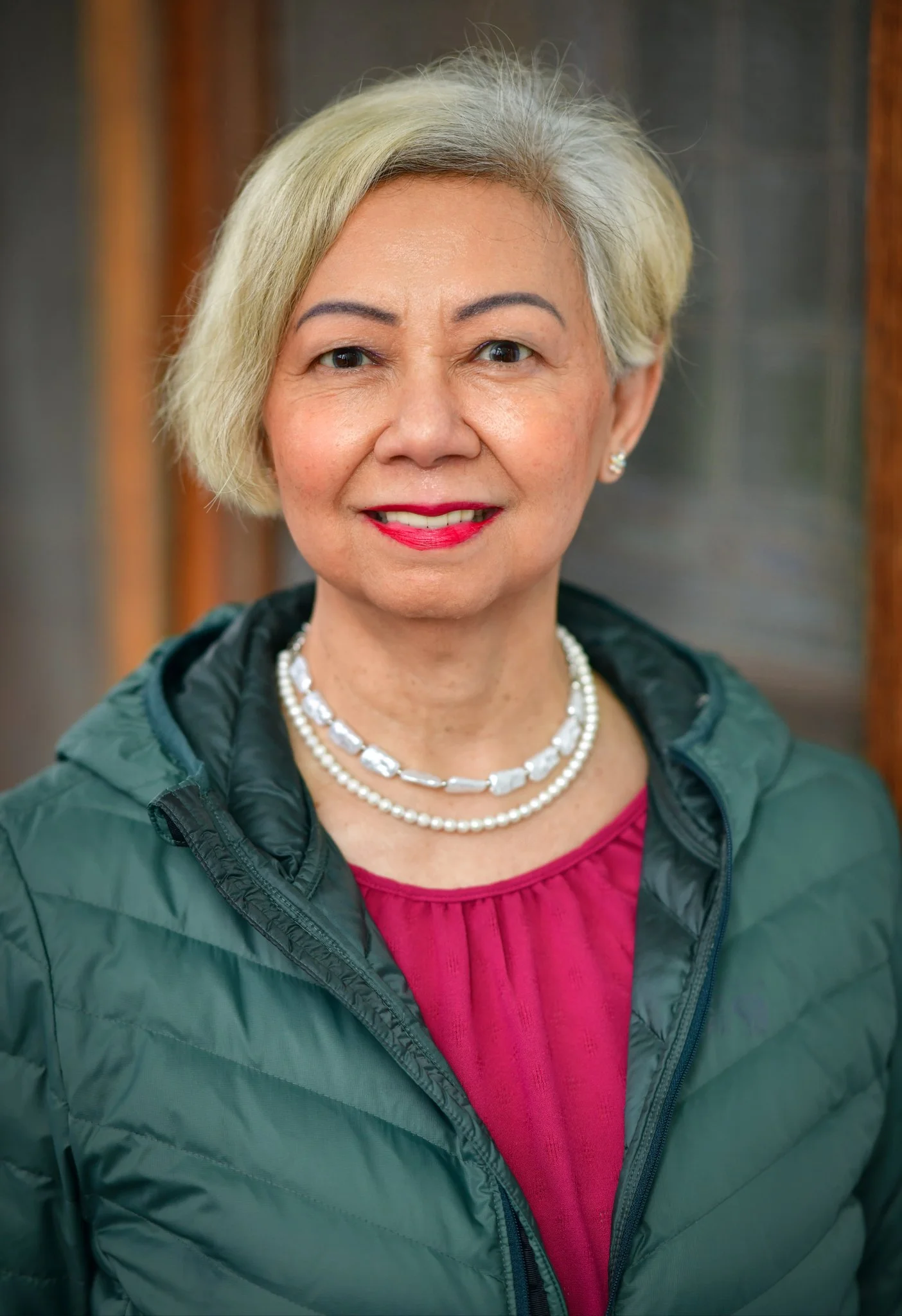 A middle-aged woman with short blonde hair smiling at the camera. She is wearing a pink top, a pearl necklace, and a dark green puffer jacket. The background is blurred with warm-colored wooden and gray elements.