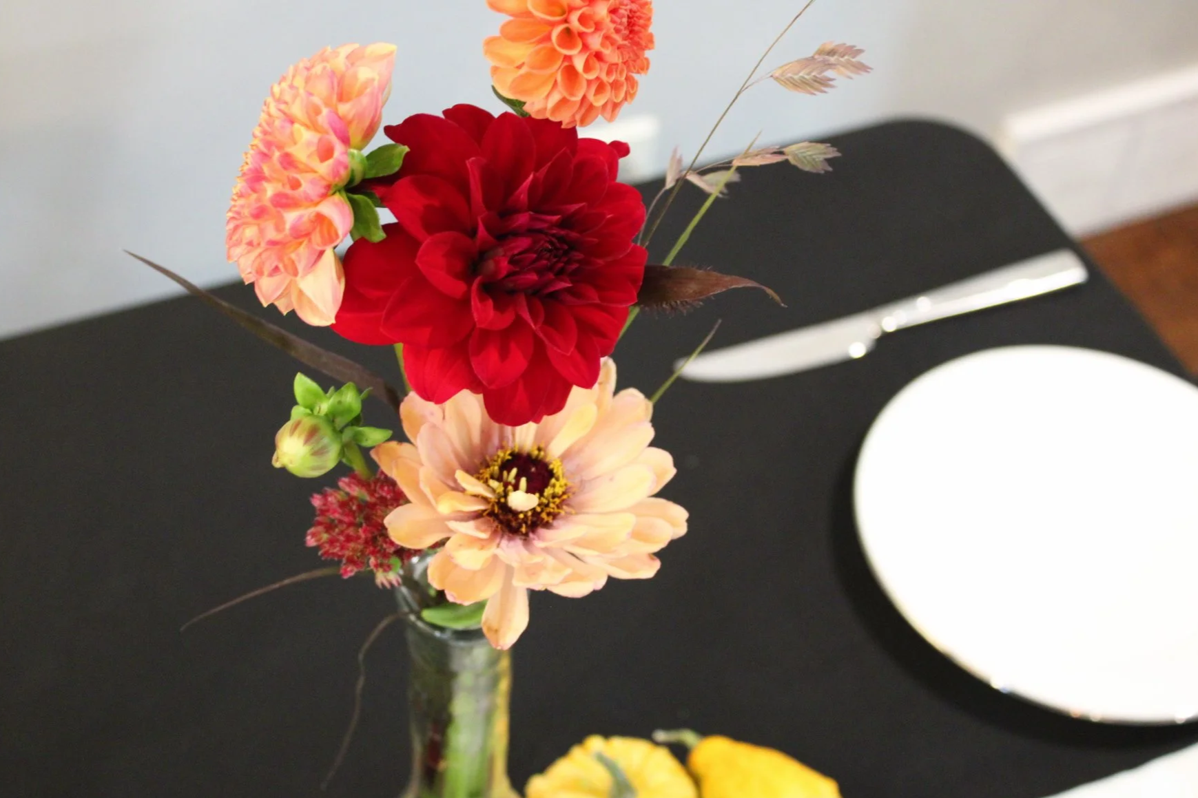 Colorful floral arrangement with red, pink, and orange dahlias in a clear glass vase on a black surface, with a white plate and fork in the background.