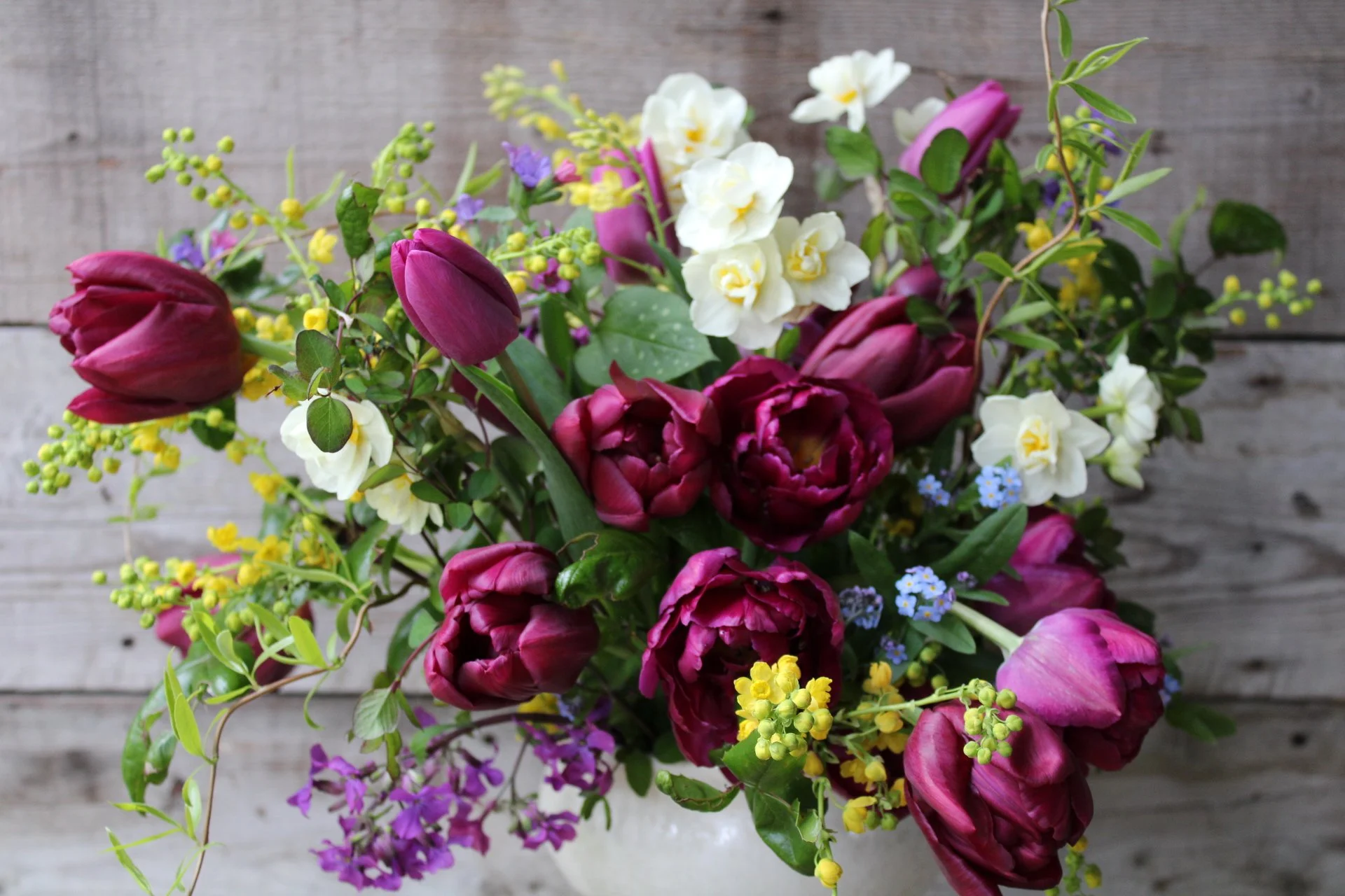 A colorful bouquet of various flowers including purple tulips, white flowers, small yellow, purple, and blue blossoms, arranged on a wooden background.