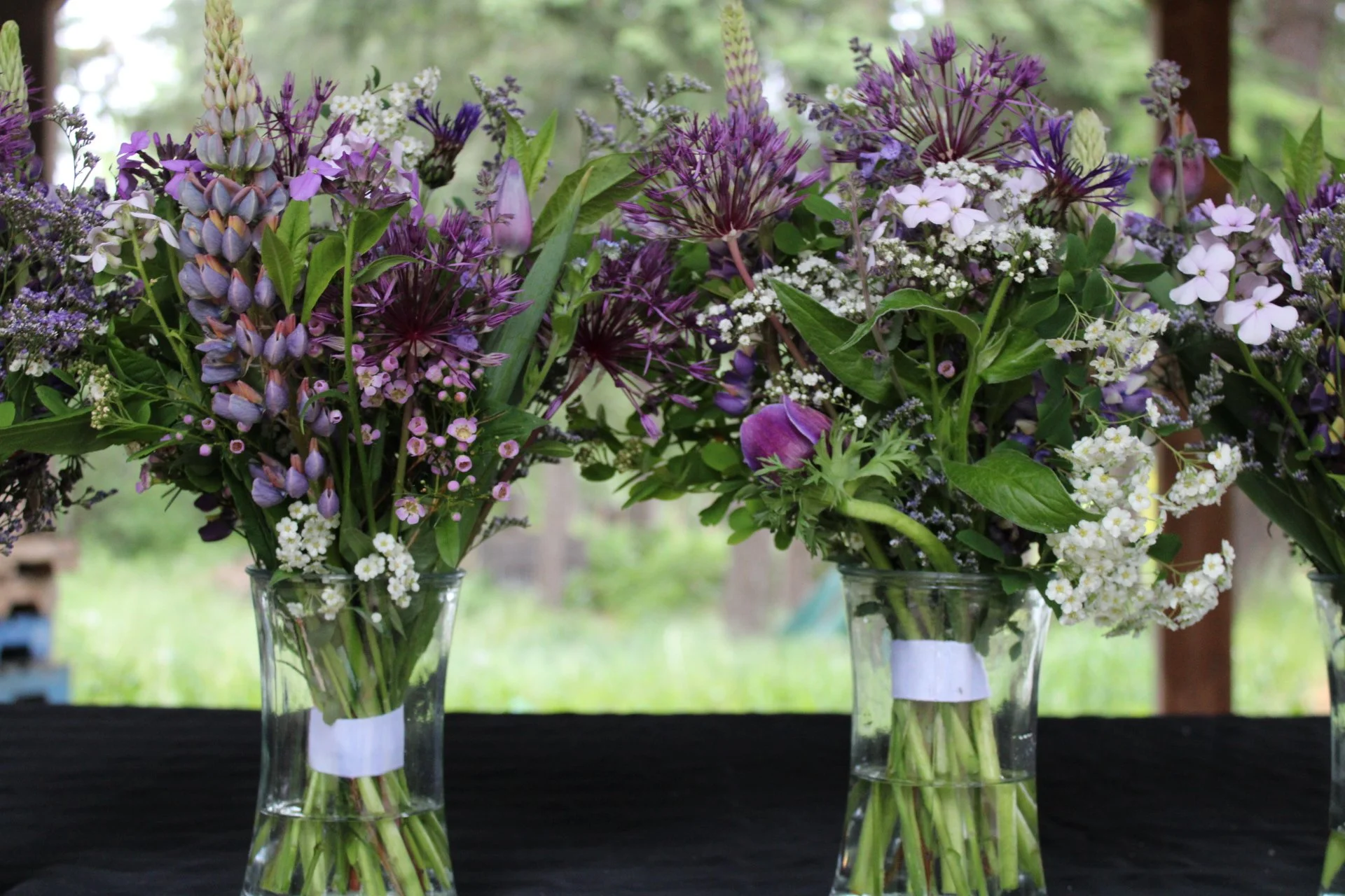 Three glass vases with colorful mixed flower bouquets, featuring purple, white, and pink flowers, placed on a black table outside with a blurred green outdoor background.