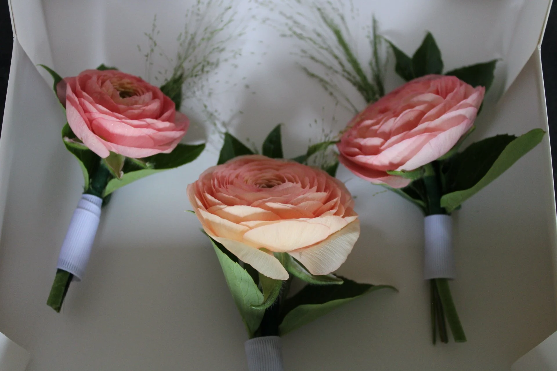 Three artificial pink and peach ranunculus flower boutonnieres with green leaves, arranged on white paper inside a box.