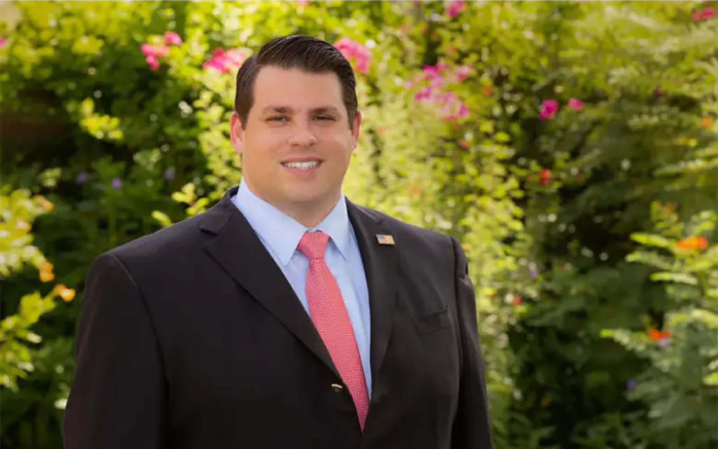 A man in a black suit and pink tie standing outdoors in front of a background of greenery and pink flowers.