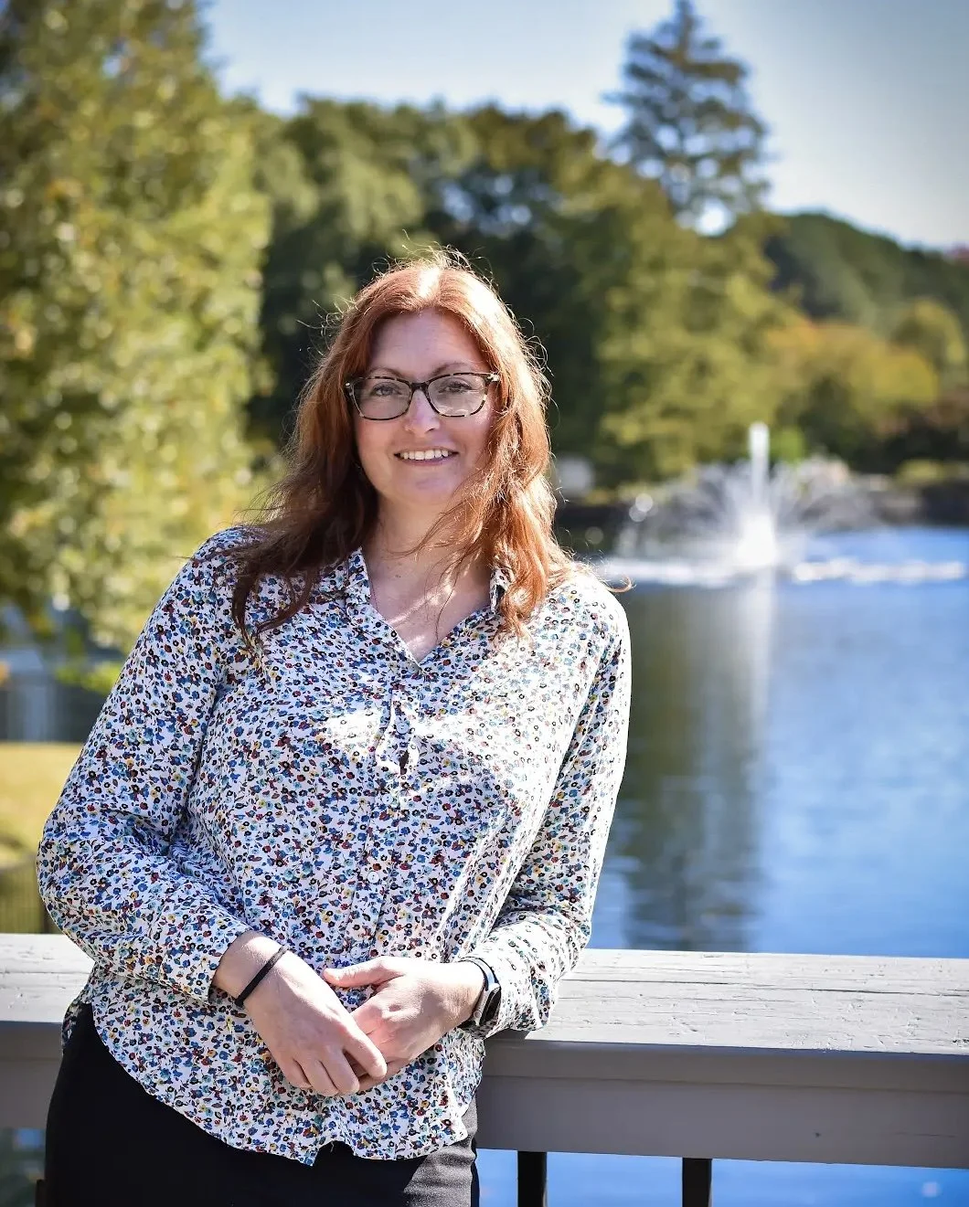A woman with red hair and glasses standing outdoors by a wooden railing, smiling, with a river and fountain in the background and trees surrounding the area.