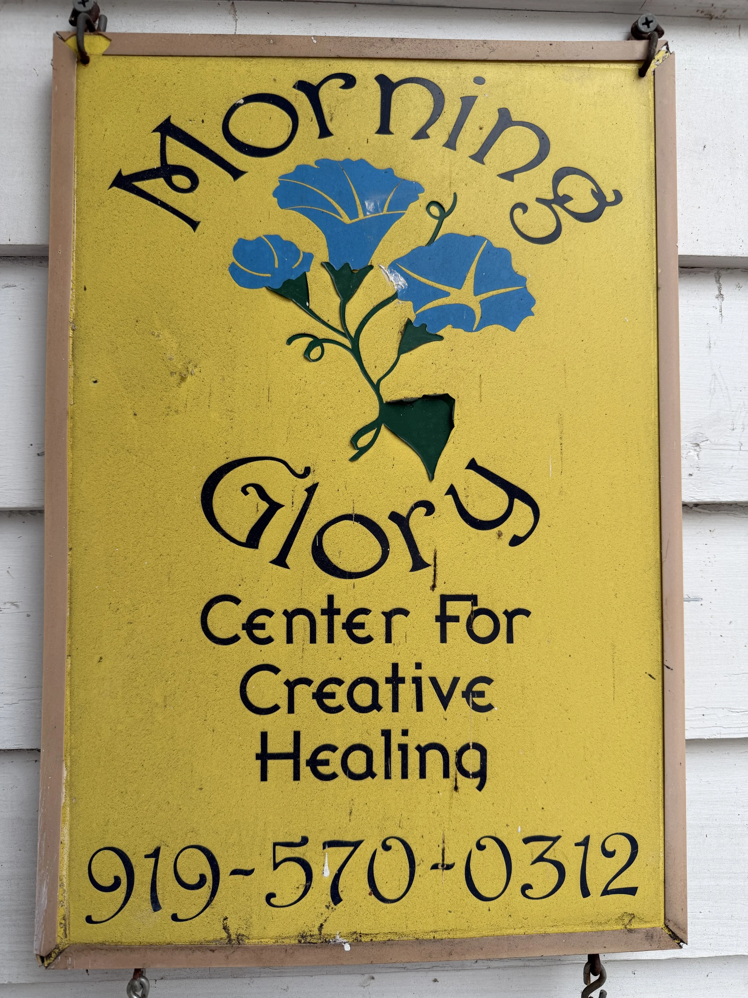 A yellow sign with black text that reads 'Morning Glory Center For Creative Healing' and a phone number, featuring a decorative blue flower design in the center.