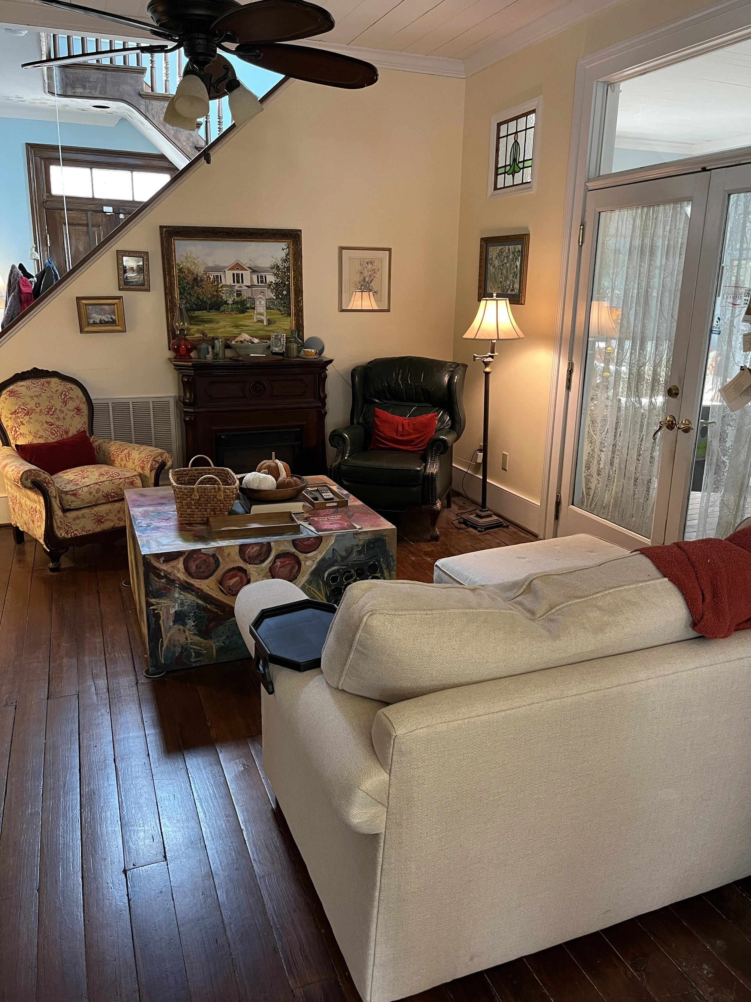 Living room with beige sofa, black leather armchair, wooden floors, artwork on walls, table with basket, and sliding glass door with lace curtain.